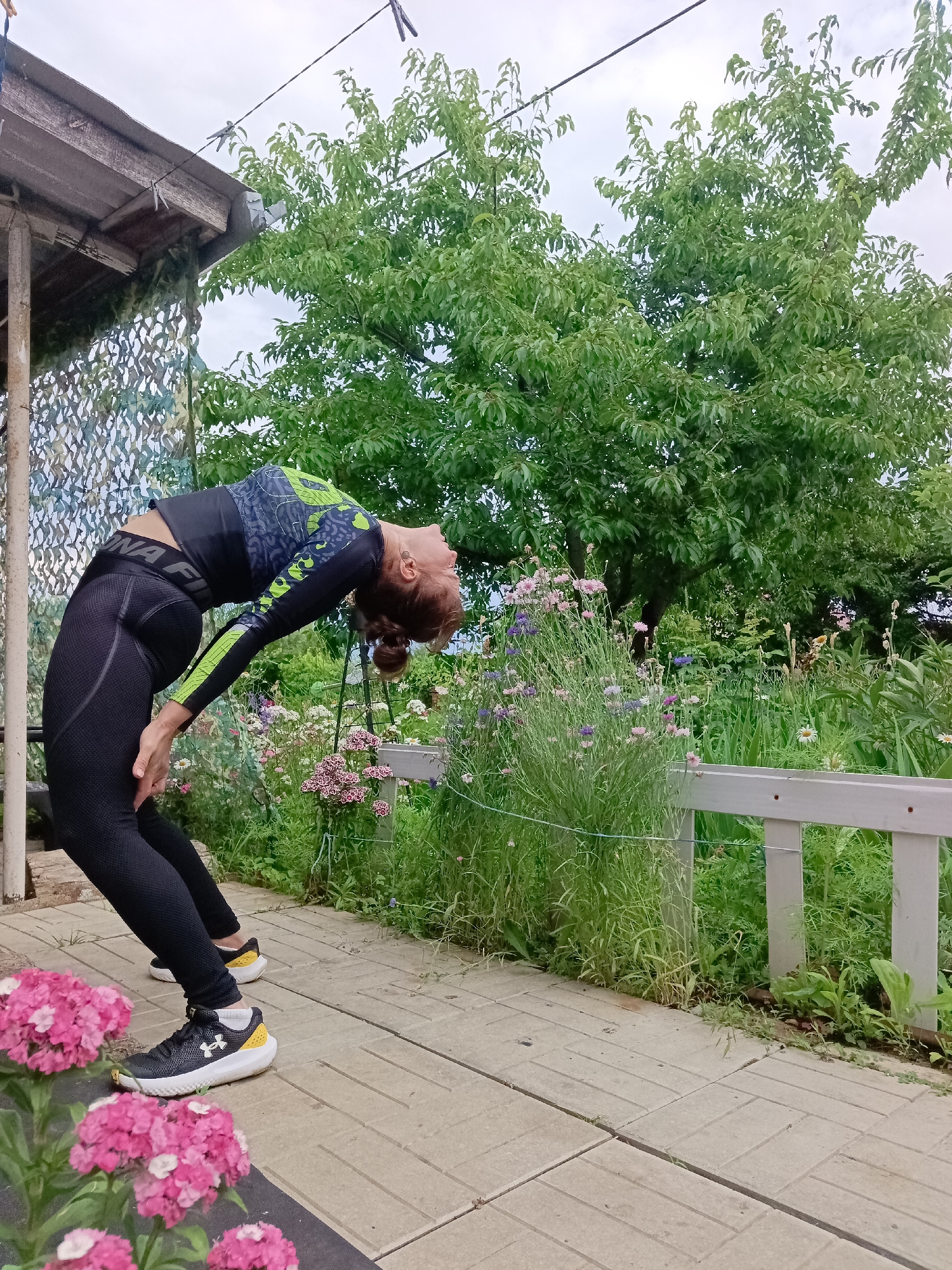 A woman with auburn hair pulled back in a bun is performing a backbend yoga pose outdoors. She is wearing a black long-sleeved athletic shirt with a green and gray pattern and black athletic leggings. Her right hand is resting on her right thigh, and her left hand is on the ground. She is on a paved walkway in what appears to be a garden or backyard. To her left is a wooden structure with a roof and camouflage netting hanging down. Behind her is a lush green tree, and in the background, there is a fence, more greenery, and flowering plants in pink, purple, and white. The sky is overcast. The location is identified as Vyazovka, Russia.