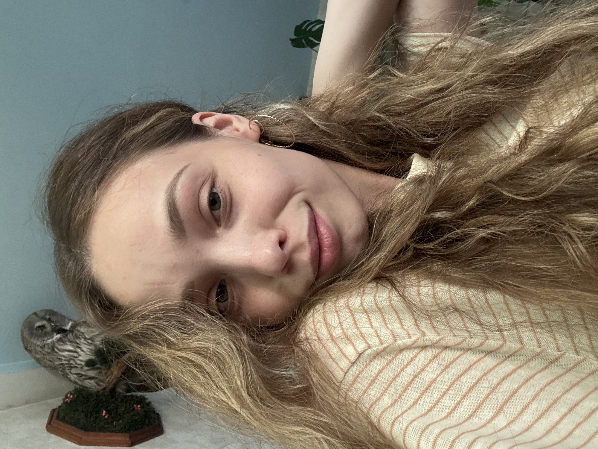 A medium shot shows a young woman with light brown, wavy hair and fair skin, looking towards the camera with a slight smile. She is wearing a light-colored, horizontally striped shirt with thin, peachy-orange lines. A large, taxidermied owl is mounted on the wall behind and to the left of the woman, perched on a brown branch against a dark wooden backing with green moss and small red decorations. The background consists of a blue wall and hints of green foliage on the right side. The woman has on a large, gold hoop earring in her right ear.