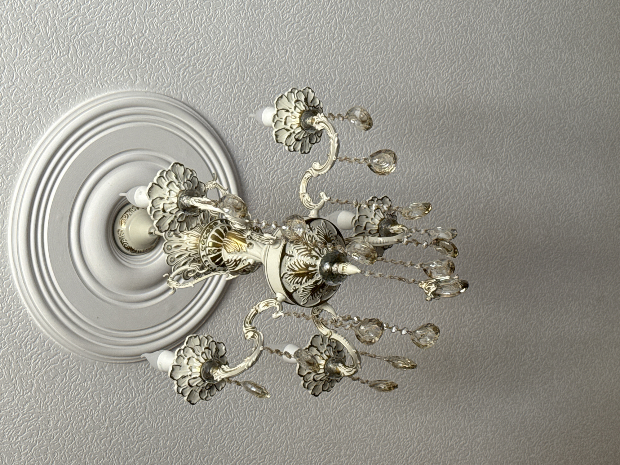 The image shows a white ornate chandelier with crystal accents hanging from a textured white ceiling with a decorative medallion. The chandelier has multiple arms, each holding a light fixture resembling a flower. The crystal pendants hang from the arms and also from the central body of the chandelier. The scene is indoors, likely in a home or a building with decorative interior features. There is no visible indication of the city of Khasavyurt, Russia.