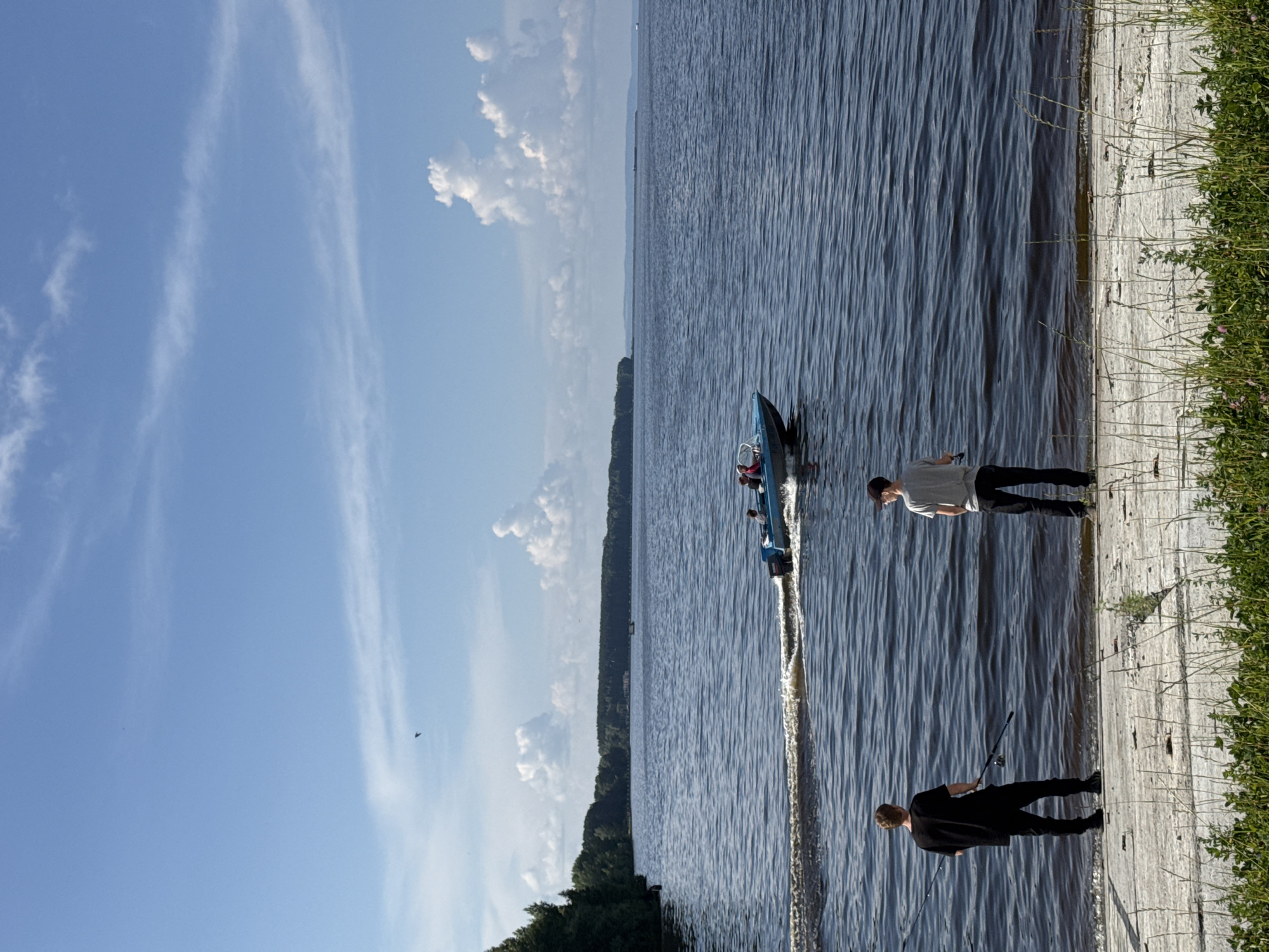 Two young men are standing on a concrete embankment beside a wide, dark blue body of water, likely a reservoir, under a bright blue sky with scattered white clouds. The man on the left, wearing a black t-shirt and black pants, holds a fishing rod. The man on the right, in a gray t-shirt and black pants, also appears to be holding a fishing rod. In the background, a small blue motorboat with several occupants is creating a wake as it moves across the water. The shoreline on the opposite side of the water is lined with a dense green forest. The foreground is dominated by the rough concrete embankment and patches of green grass and some pink wildflowers. The scene appears to be a serene outdoor activity in a natural setting, possibly on the Kama River near Chaikovsky, Russia, given the characteristic wide river and forest landscape.