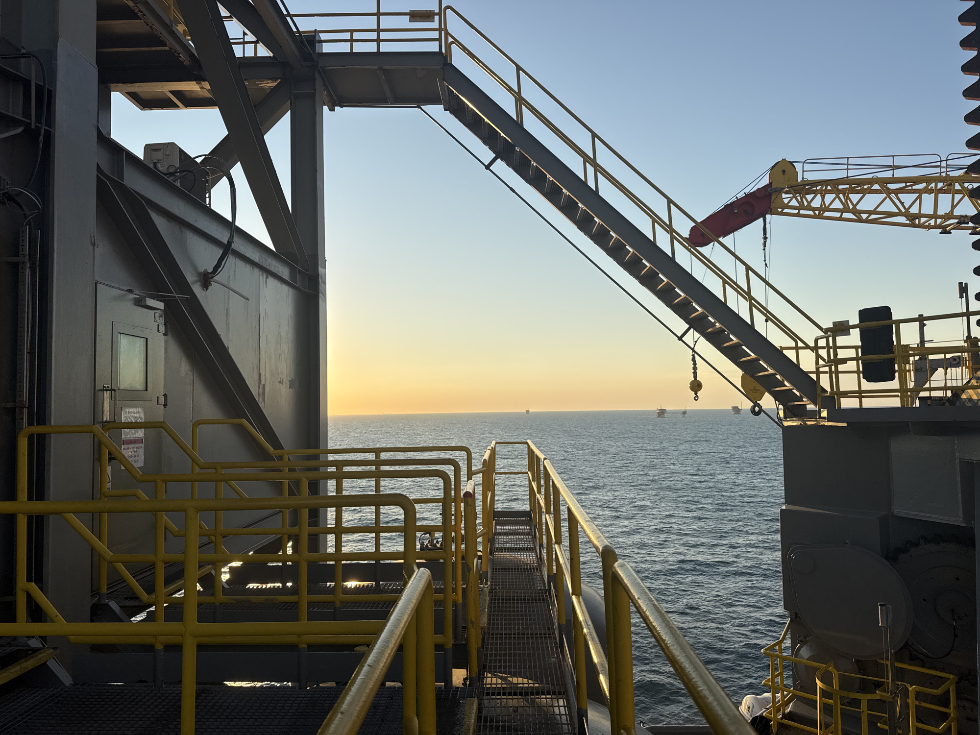 The image depicts an industrial scene on an offshore platform at sunrise. The foreground features yellow metal walkways and staircases, with safety railings. A large, dark gray industrial structure dominates the left side of the frame, with visible conduits and a door with a window. To the right, a yellow crane arm extends across the scene, with a red component hanging from it. The background opens up to a calm, blue sea under a clear sky transitioning from pale yellow near the horizon to a light blue above. Several distant oil rigs or similar structures are visible on the horizon. The lighting suggests either early morning or late afternoon. The scene is devoid of any people. The location is not in the city of Unknown, Italy; it is an offshore industrial setting.