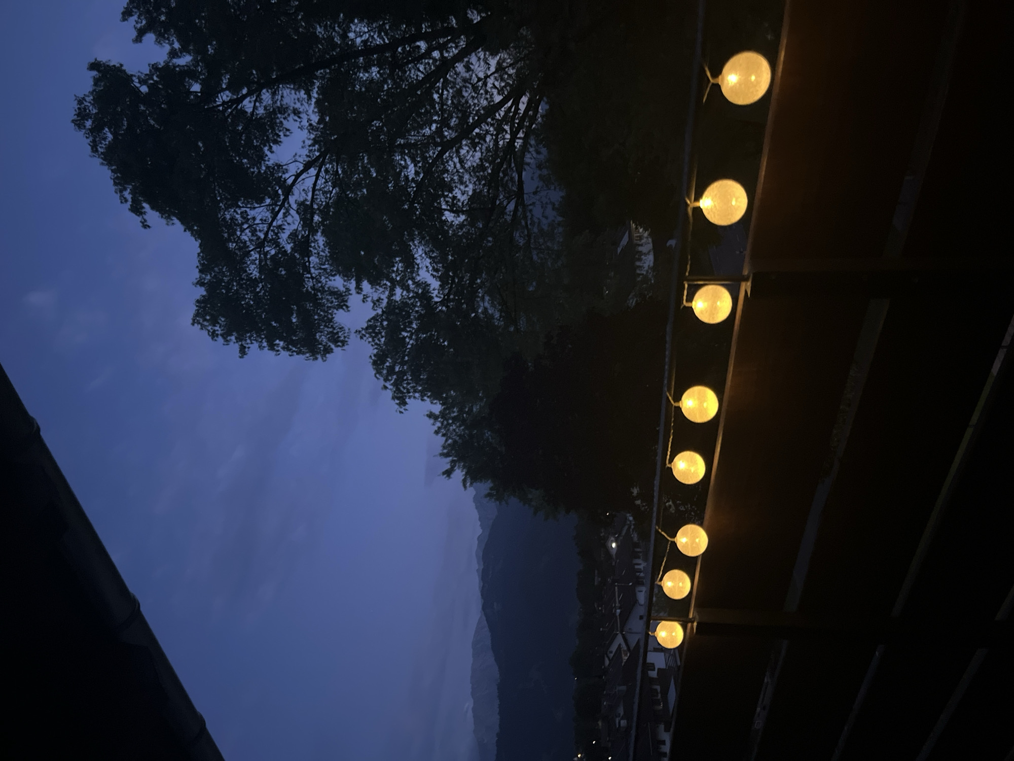 The image shows a string of illuminated globe lights across a dark balcony railing. In the background, silhouetted trees and distant mountains are visible under a deep blue twilight sky. The lights cast a warm glow on the railing. Below the railing, hints of houses and a town can be discerned. The scene suggests an evening view from a balcony in a mountainous region. Given the context of Garmisch-Partenkirchen, Germany, this is likely a view overlooking the town and the surrounding Alps.