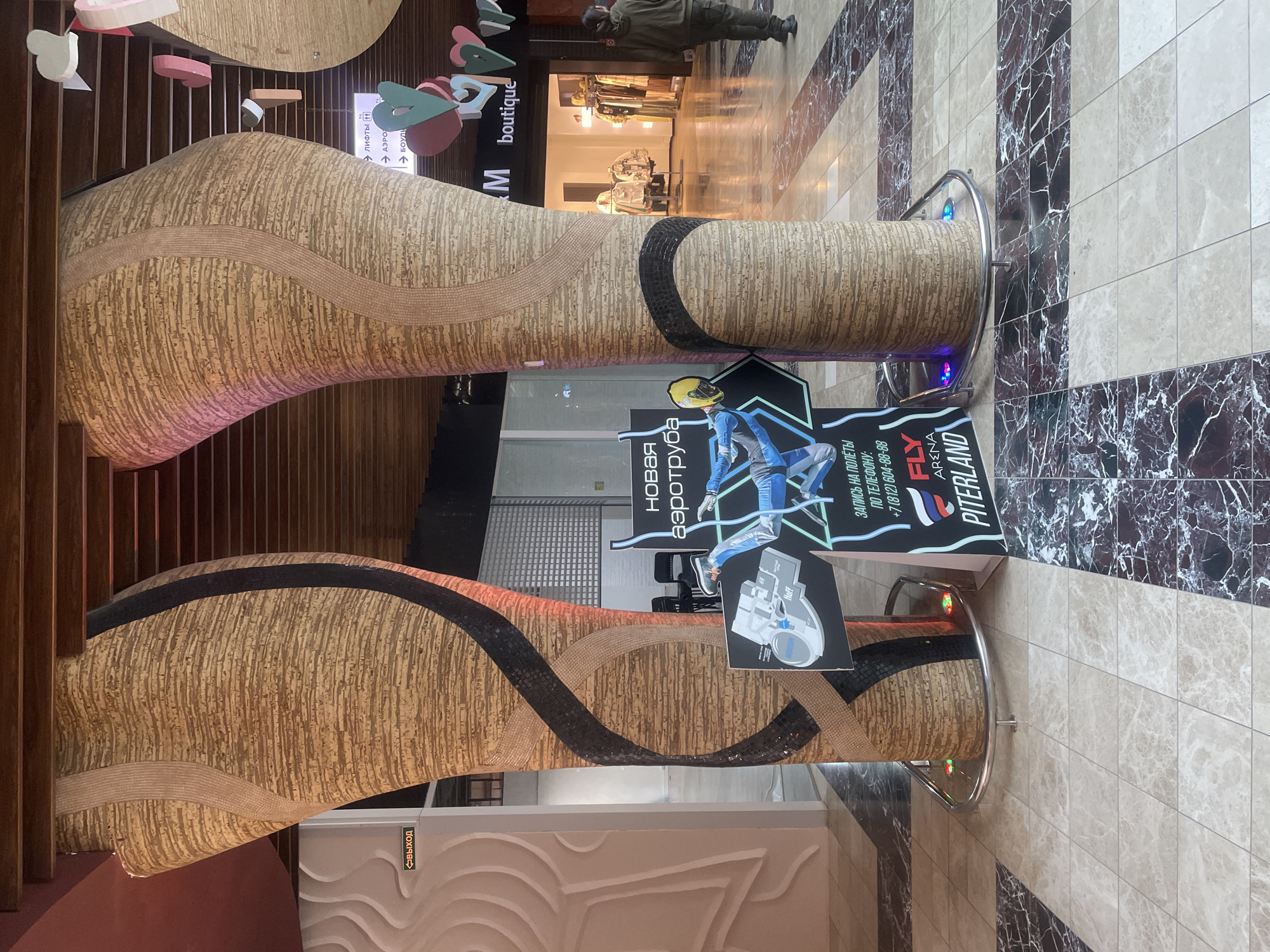 In the city of Saint Petersburg, Russia, the image depicts a section of an indoor shopping mall with a distinctive architectural design. Two large, curved pillars with a textured, cork-like surface and inlaid wavy bands of black and light brown mosaic dominate the foreground. The pillars are illuminated with subtle colored lights at their base. In front of one pillar, a sign advertises "NOVA AEROTRUBA" (New Aerotube) with an illustration of a person in mid-air, suggesting an indoor skydiving attraction. The sign also provides a phone number. Further into the mall, a boutique store with clothing displays is visible. A person in a dark green hooded jacket and pants is walking away from the camera towards the right. The mall floor is tiled with a pattern of beige and dark brown marble. A sign with an arrow and the word "ВЫХОД" (Exit) is mounted on a white wall to the left. Decorative heart shapes are suspended from the ceiling in the background. The environment suggests a modern commercial space designed for entertainment and retail.