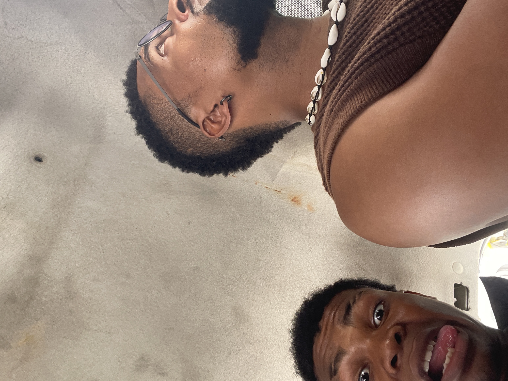 Two dark-skinned young men are positioned close to the camera in an indoor environment with a light gray background. The man in the upper part of the frame is wearing eyeglasses, a brown sleeveless shirt, and a beaded necklace, and has a neatly trimmed beard and mustache. The man in the lower part of the frame is sticking his tongue out, facing the camera, and has short hair. The photograph appears to have been taken from an overhead angle. The location and city are not discernable from the provided image.
