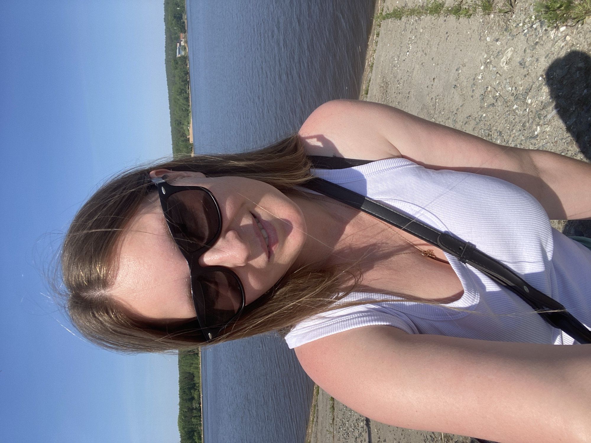 A woman in sunglasses takes a selfie outdoors in sunny conditions. The background shows a body of water, possibly the Volga River, with forested land visible on the opposite shore. She is standing on a concrete surface with sparse vegetation. She is wearing a white shirt and a black strap of some sort crosses her torso. The location is likely near Noviy, Russia, given the Volga River presence.
