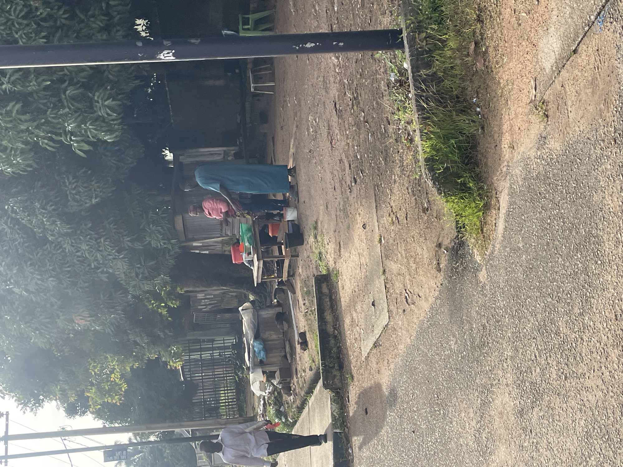 A medium shot shows a street scene in Jos, Nigeria, with a man in a white shirt and dark pants walking from left to right on a sidewalk. Behind him, a woman in a pink top and dark pants and a person in a teal robe stand near a table laden with various items, including buckets, bowls, and what appears to be produce. The ground is a mix of dirt and gravel. Trees with lush green foliage are visible in the background, along with a metal fence and what looks like a market stall. A dark lamppost stands on the right side of the frame. The lighting suggests it is daytime, with sunlight casting shadows.