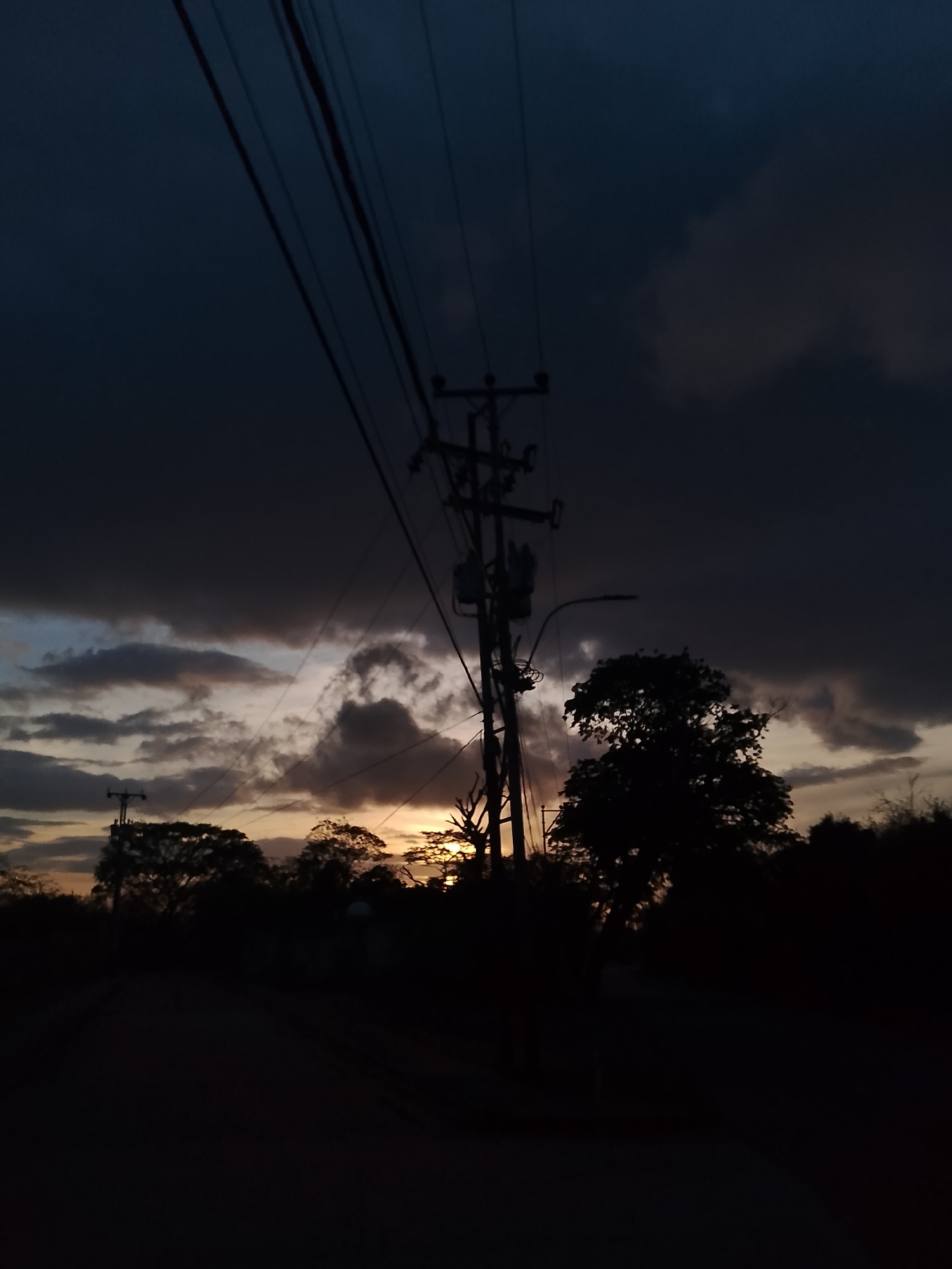 A silhouette of a utility pole with electrical wires and transformers stands against a twilight sky, obscuring much of the detail in the foreground and midground. Dark, heavy clouds dominate the upper portion of the sky, with lighter, textured clouds visible towards the horizon where a hint of sunset orange can be seen. Silhouetted trees and vegetation are scattered across the lower part of the image, with some appearing denser and taller than others. The ground in the foreground is dark and indistinct, suggesting it might be a road or open area. The scene is devoid of human figures or any discernible activity. The overall impression is one of a rural or semi-rural setting at dusk.