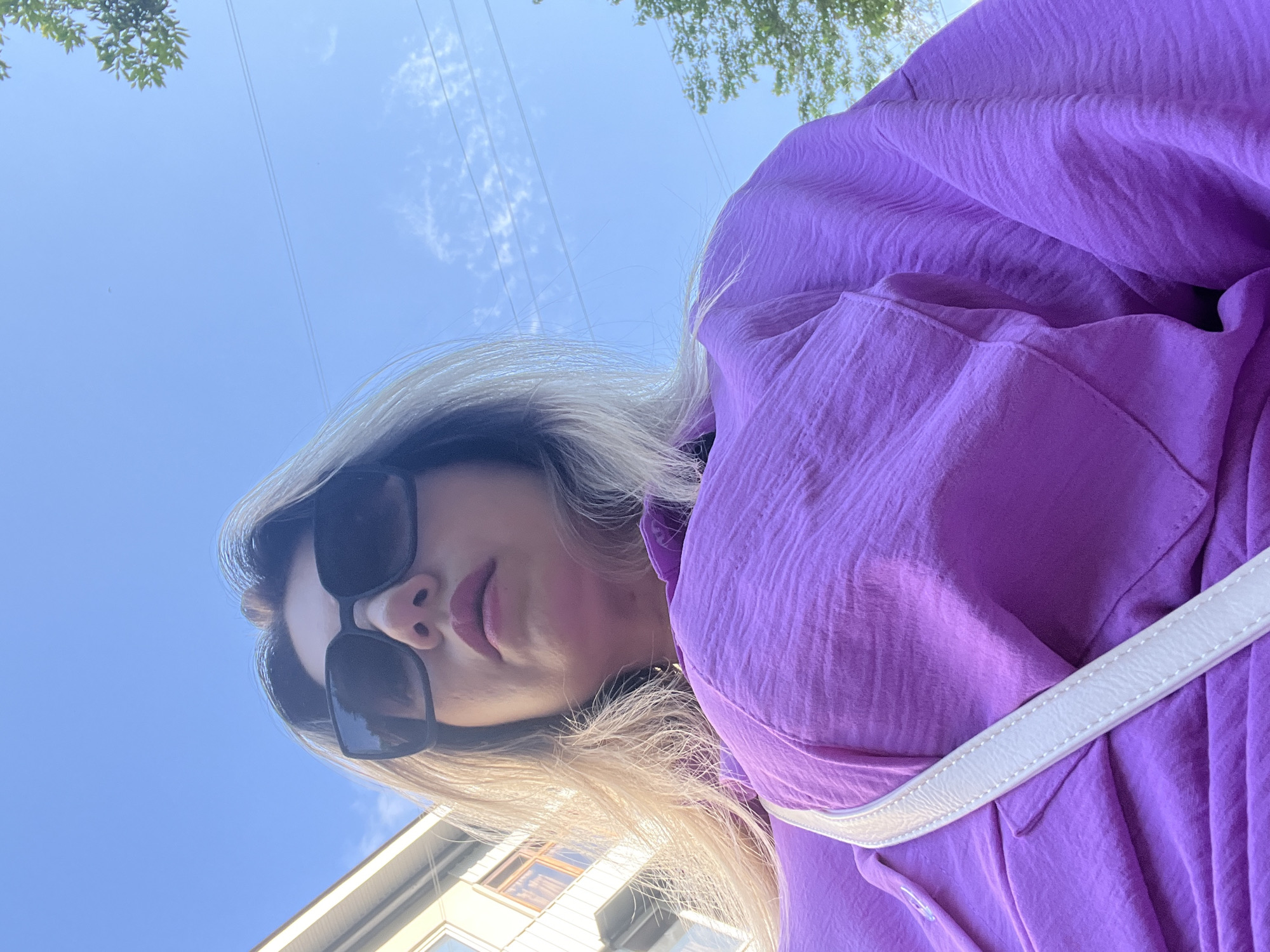A woman is captured in a low-angle, close-up shot, looking upwards as if at the camera. She is wearing dark sunglasses and a vibrant purple top that has a textured, crinkled fabric. A thin white strap, likely from a bag, crosses diagonally over the fabric. Her blonde hair falls around her face. The background is a bright blue sky with a few wispy white clouds, suggesting a clear day. Tree branches are visible at the top of the frame, and a hint of a building with windows can be seen at the bottom left. The sunlight casts a warm glow on her face and hair. The overall impression is of a casual, outdoor moment.