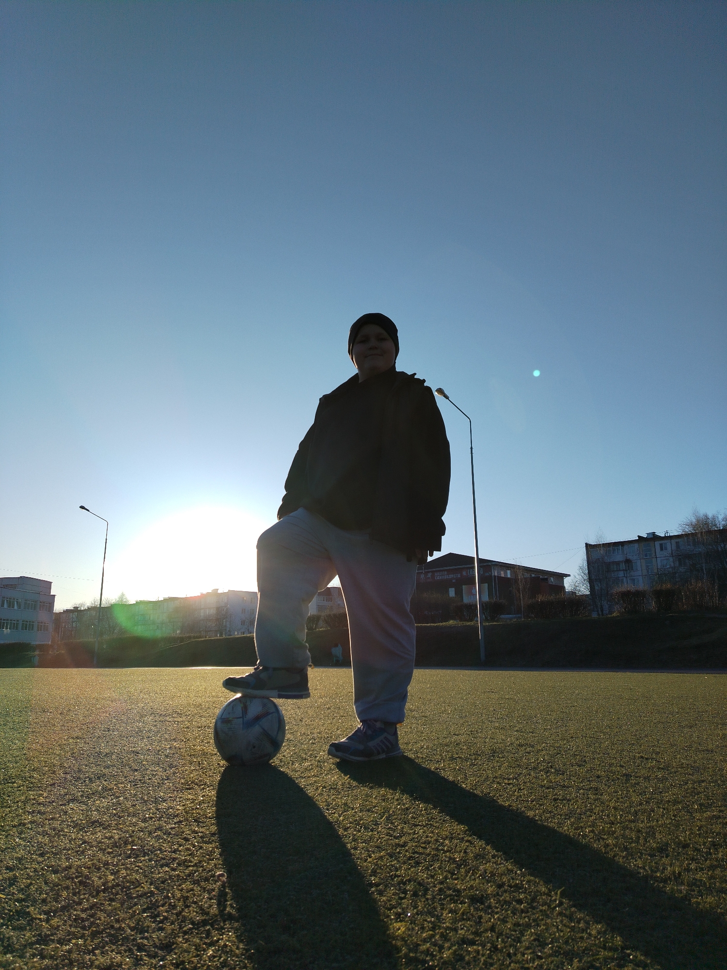 A person in a dark jacket, light gray pants, and a dark beanie stands on a soccer ball on a green artificial turf field. The person's right foot is resting on the ball. The person is facing the camera, with a visible shadow cast to the right. The background consists of a clear, light blue sky and buildings with trees, illuminated by the setting sun which is situated in the top left quadrant of the picture. Streetlights are also visible behind the person. Based on visual indicators such as building architecture and general landscape features, the image appears to have been taken in the city of Chaikovsky, Russia.