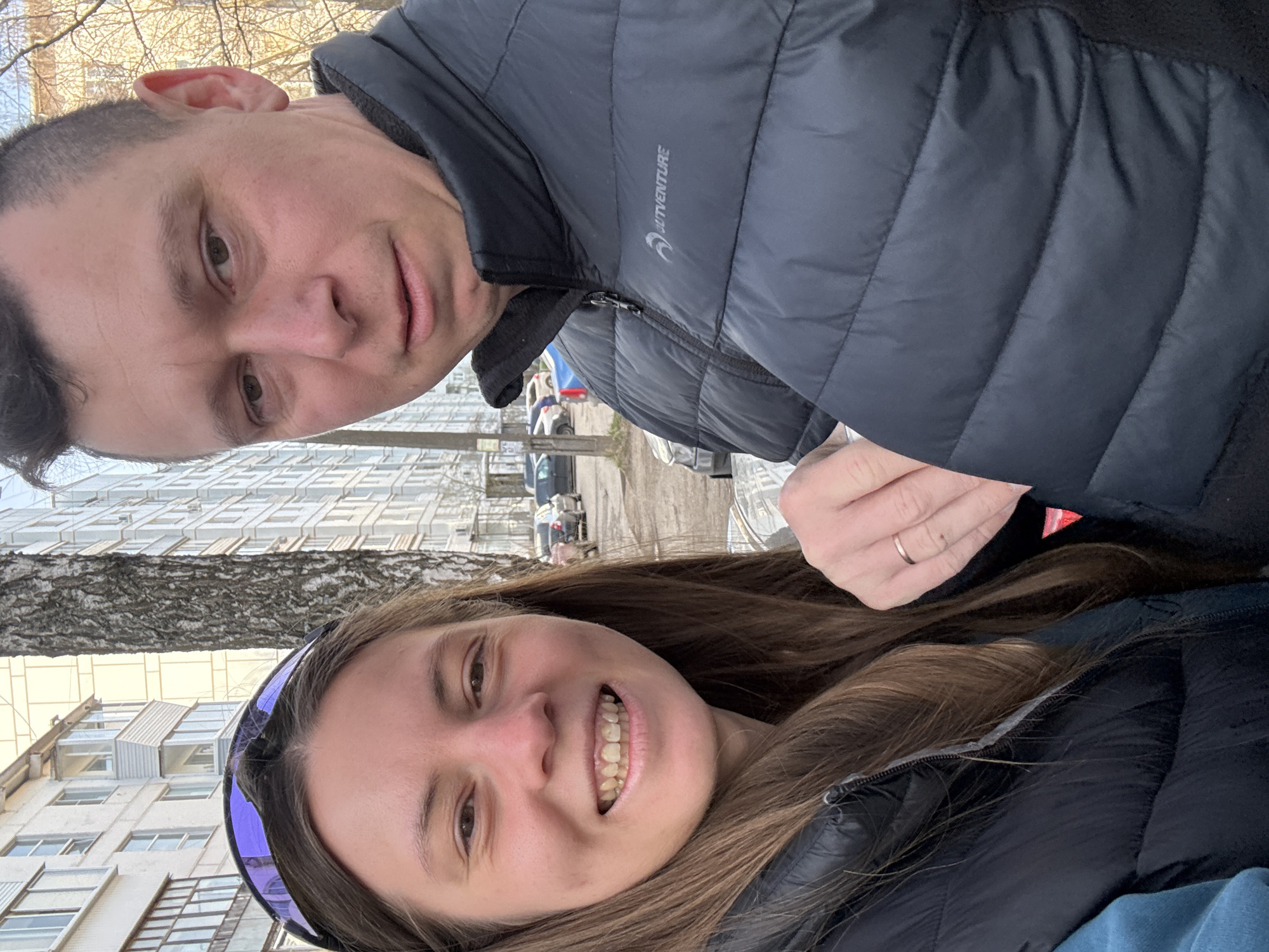 A vertical, slightly rotated selfie features two individuals, a man and a woman, positioned in the foreground. The man, with short dark hair and a neutral expression, occupies the upper part of the frame. He wears a dark grey or black puffer jacket with a visible logo and the text "OUTVENTURE" on the chest. His ear is visible, and he has a slight stubble.
Below and slightly in front of him is a woman with long brown hair, smiling broadly and showing her teeth. She wears a dark puffer jacket similar to the man's and a purple headband pushed up into her hair. A simple gold-colored ring is visible on her left hand.
The background depicts an urban setting, consistent with Chaikovsky, Russia. Dominant features include multi-story residential buildings with numerous windows, suggesting apartment complexes. A large tree with bare branches, indicative of late winter or early spring, stands between the subjects and the buildings. In the further distance, what appears to be a street with parked cars and other structures can be seen. The lighting suggests it is daytime, with diffused light possibly from an overcast sky or an early morning/late afternoon sun. The warm clothing worn by both individuals further supports a cooler season.