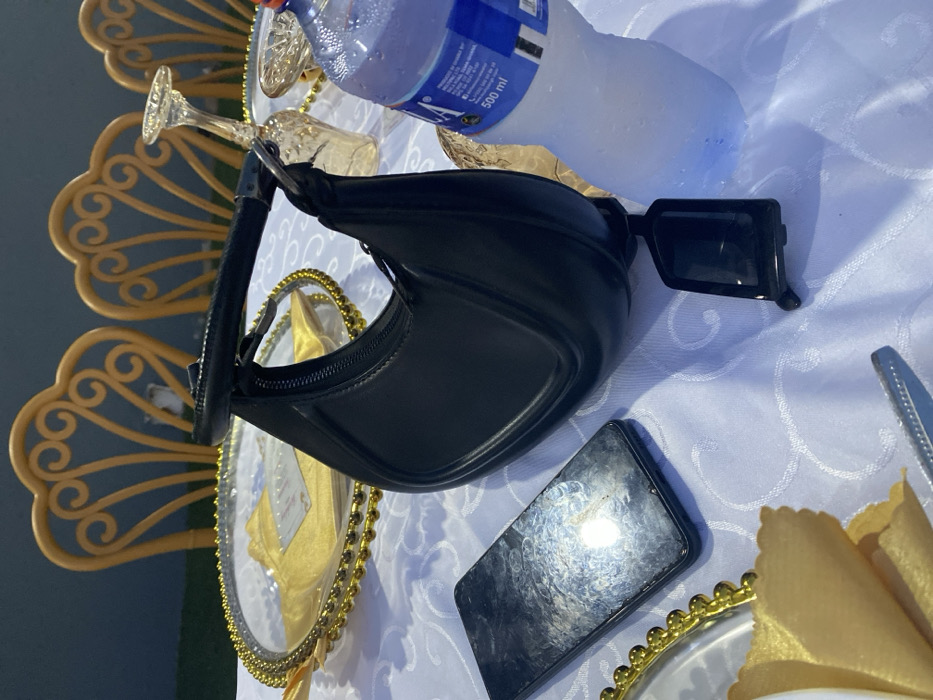 A black purse, a smartphone with a mirrored screen, and black sunglasses rest on a white tablecloth next to a water bottle. A golden decorative plate and a glass with a stem can also be seen on the table. A golden chair with a fan design is slightly visible in the background.
