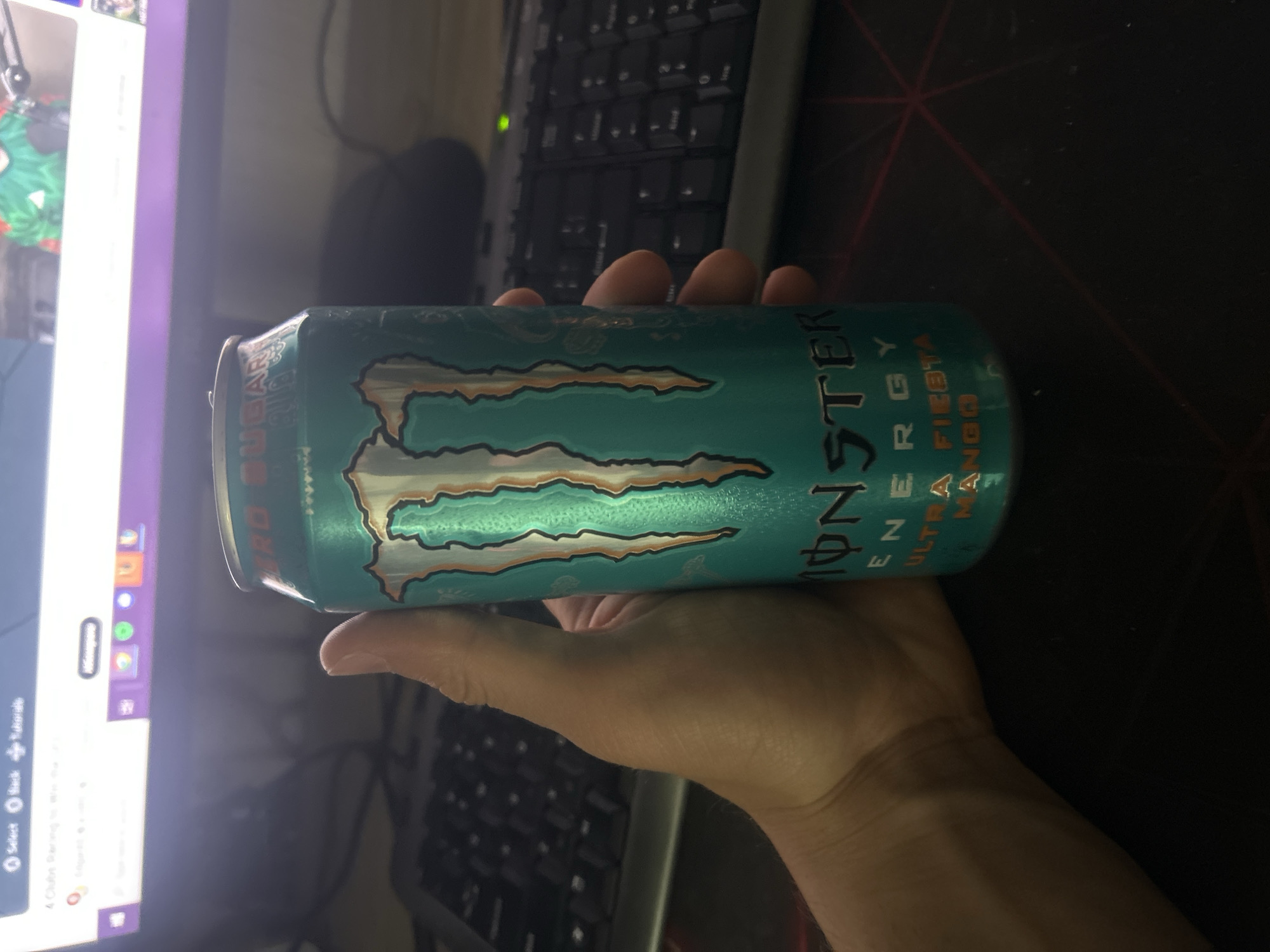 A person's hand is holding a teal-colored can of Monster Energy Ultra Fiesta Mango drink. The can is positioned horizontally. A computer monitor showing a webpage is visible in the background. A keyboard and mouse pad are also partially visible in the background. The location is not identifiable as Elhovo, Bulgaria from the provided image.