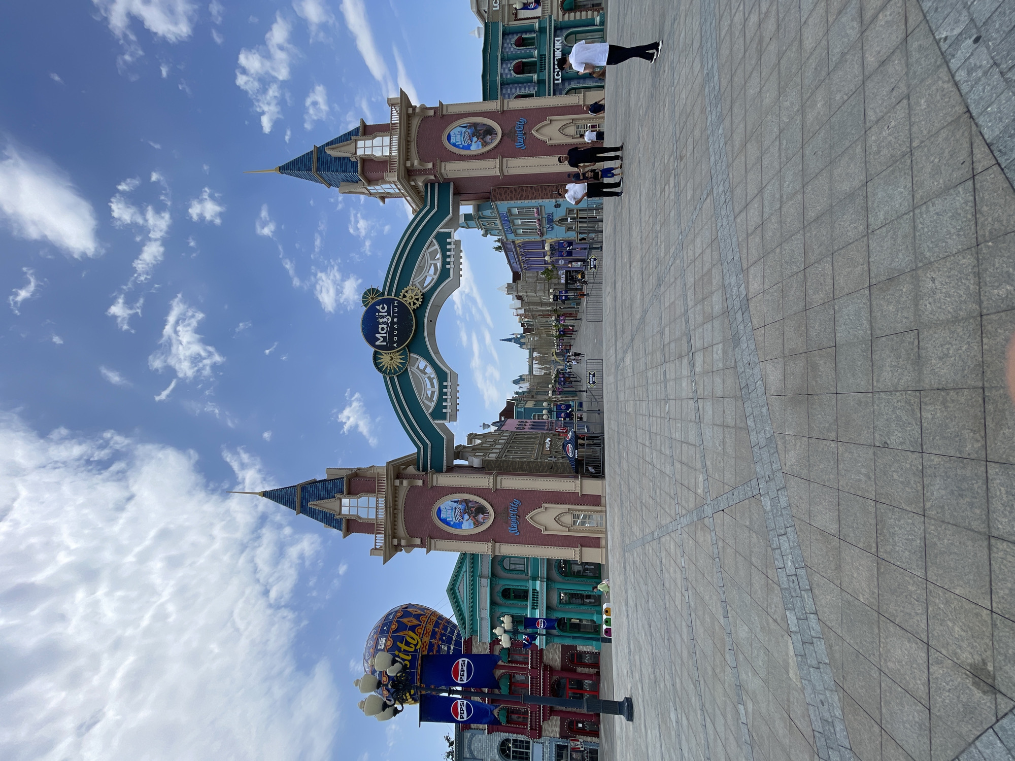 A wide-angle, eye-level shot captures a spacious, paved plaza in front of an amusement park entrance. The plaza is constructed with large, square grey tiles arranged in a grid pattern with inlaid granite strips forming diagonal and perpendicular lines. In the background, a large, archway-like structure marks the entrance to "Magic City Aquarium" in Tashkent, Uzbekistan. The arch is adorned with teal and gold detailing and topped with two castle-like towers. The towers have red brick bases and blue roofs with spires. On either side of the entrance, colorful buildings with elaborate facades line a pedestrian walkway leading further into the park. A hot air balloon is visible to the left, suspended above a lamppost adorned with Pepsi logos. Several people are present in the scene: a family of three (two adults and a child) stand near the archway, a man in a white shirt and black pants walks away from the camera towards the right, and other individuals are scattered throughout the background, some walking and others standing. The sky above is a vibrant blue with scattered white clouds. The overall atmosphere appears bright and sunny, typical of a daytime scene in a public entertainment venue.