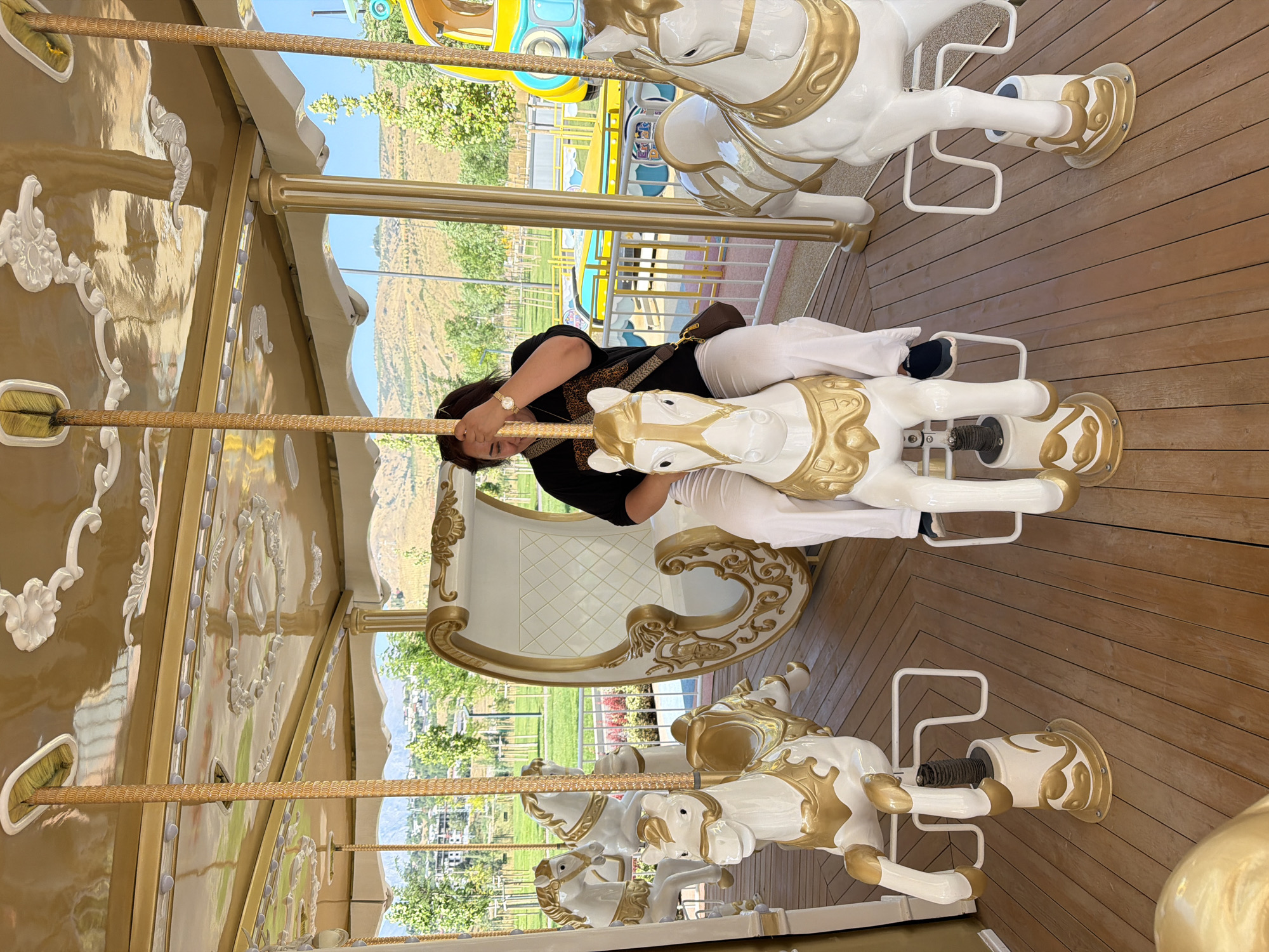 In Chinarkent, Uzbekistan, a woman sits on a white carousel horse with gold accents. She wears a black t-shirt and white pants. The carousel, adorned with ornate gold trim and detailed carvings on its ceiling, is set against a backdrop of a park with trees and rolling hills. Other carousel horses and a decorative carriage are visible. The ground of the carousel is made of polished wooden planks. The sky is a clear blue, suggesting a sunny day. In the background, part of a yellow and blue amusement park ride is visible.