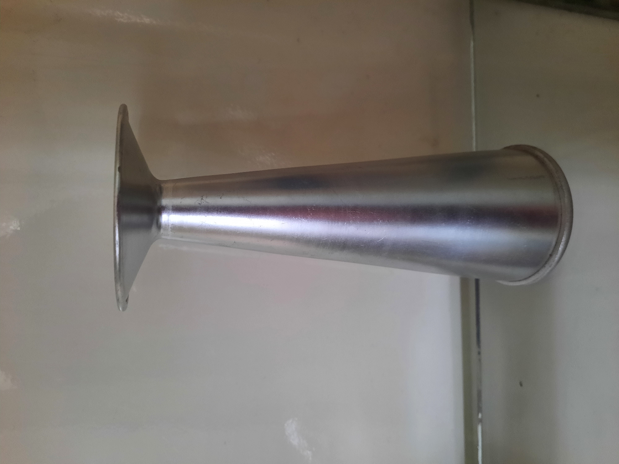 A metallic funnel-shaped object, possibly made of aluminum, stands upright on a flat surface. The object is silver in color and appears to be smooth. The wider opening is at the top, flaring outwards, while the bottom tapers to a narrower opening. The background is a plain, light-colored surface, possibly a wall or a shelf, with some subtle vertical lines or texture. The lighting is diffused, creating some reflections on the metallic surface of the object. There are no people or other objects visible in the image. The object is positioned in the center of the frame. The location in Lhokseumawe, Indonesia is not evident from the visual information provided in the image.