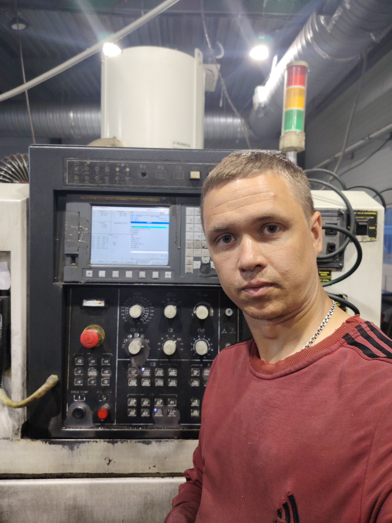 A man stands in front of a CNC (Computer Numerical Control) machine. The machine has a control panel with a screen displaying lines of code, several knobs, and a red emergency stop button. A light tower with red, yellow, and green lights is visible in the background. The man is wearing a maroon long-sleeved shirt with black stripes on the sleeves and a silver chain. The environment appears to be a manufacturing or industrial setting. The location is not discernible as Noviy, Russia from the information presented.