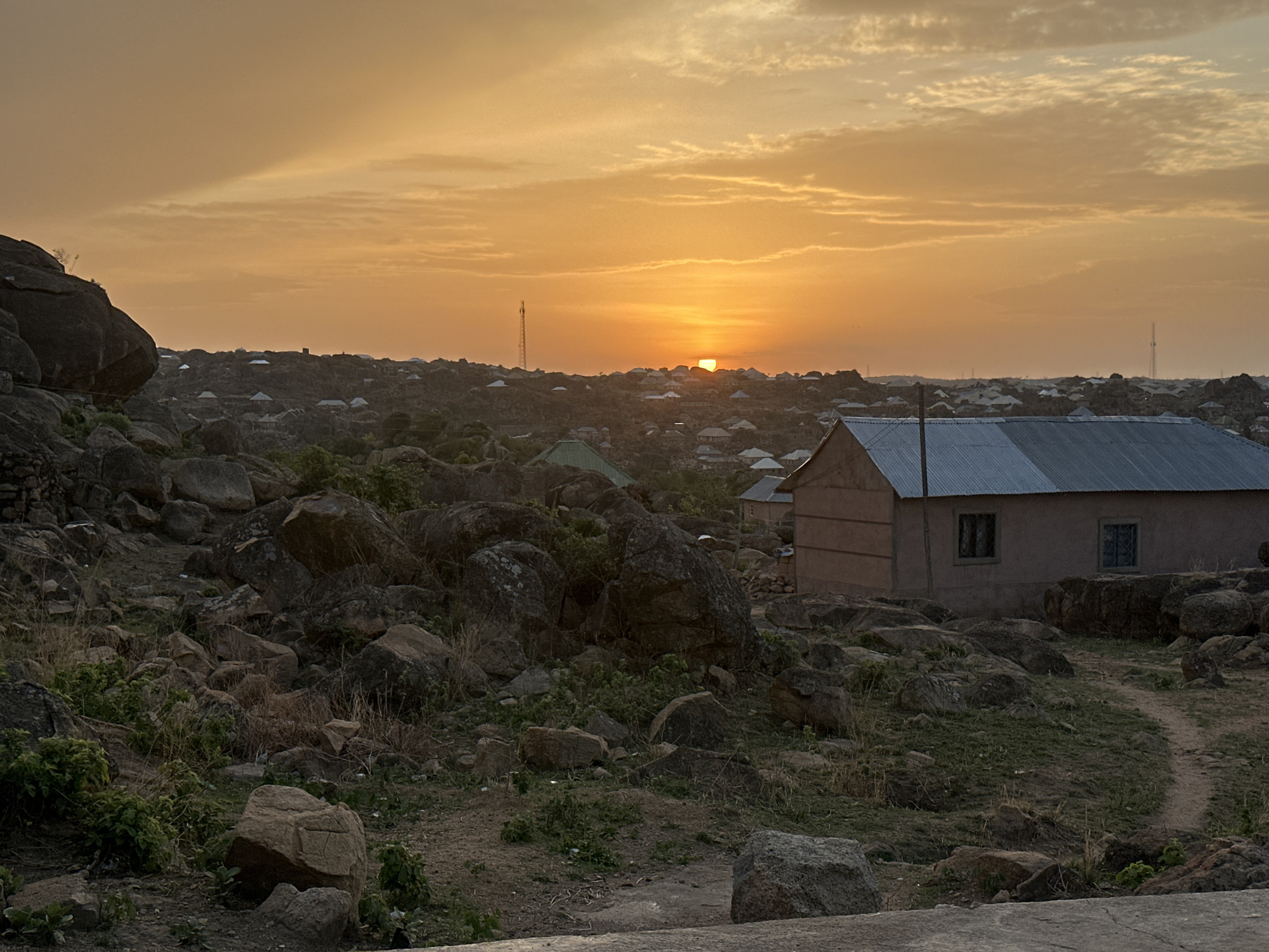 The image depicts a sunset view in Kabong, Nigeria, showcasing a landscape dominated by rocky terrain and interspersed with sparse vegetation. A small, single-story building with a metal roof is situated in the foreground to the right. In the distance, numerous rooftops suggest a densely populated area nestled amidst the rocky outcrops. The setting sun is partially obscured by the horizon and casts an orange glow across the sky and the scene. A communication tower is visible on the horizon to the right of the sun. No people are visible in the image.