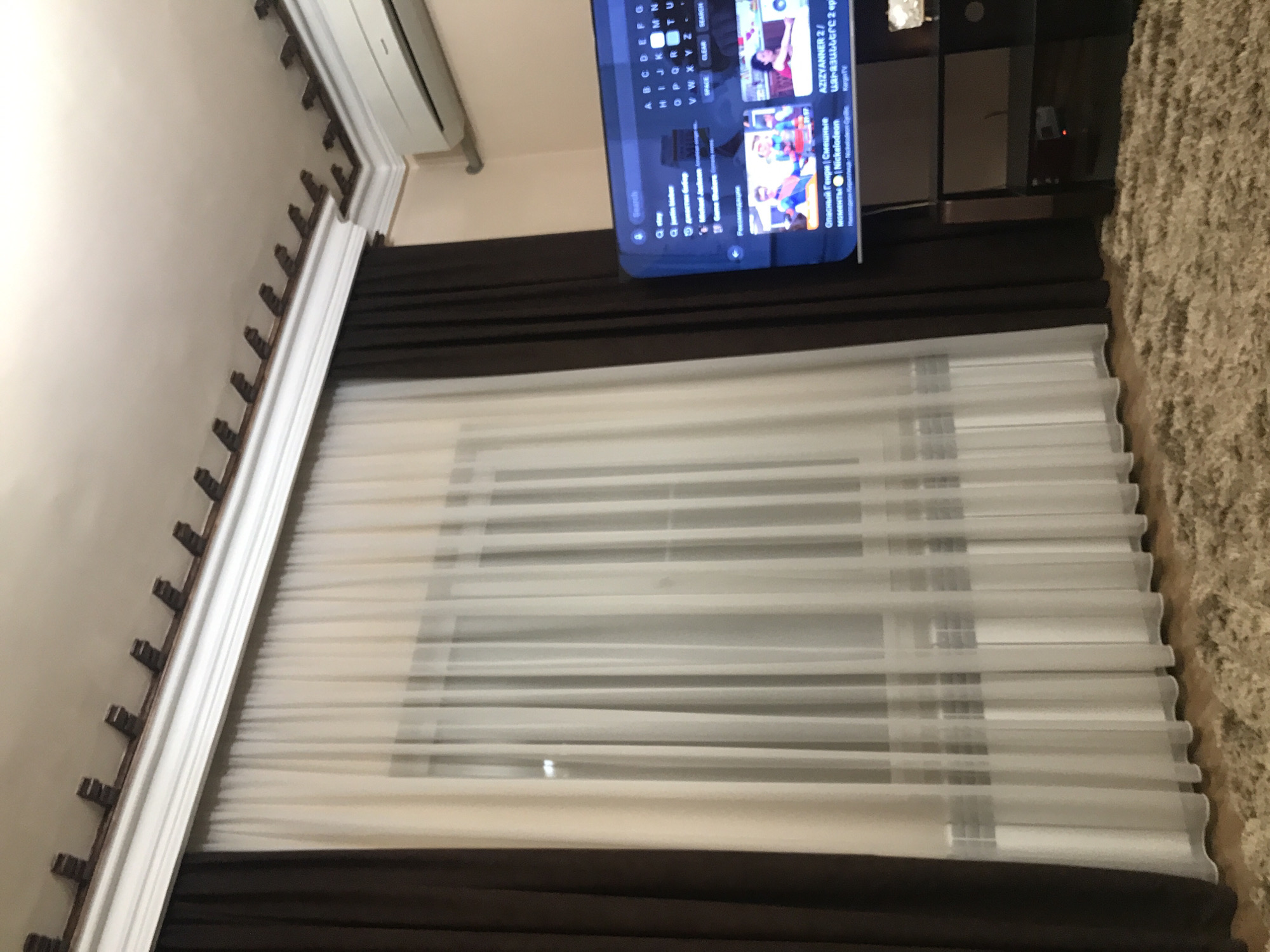 The image shows a window with layered curtains and blinds, above which a television is mounted. The top and bottom curtains are a dark brown, and the middle layer consists of sheer, white blinds. The television screen displays what appears to be a streaming service menu, with Armenian and Russian text visible, suggesting location in Yerevan, Armenia. A section of a white crown molding is visible on the left side of the frame. A dark-colored shelf or cabinet is to the upper right of the television, and a textured, possibly carpeted surface is visible on the right side of the image. An air conditioning unit is seen in the upper left of the image.