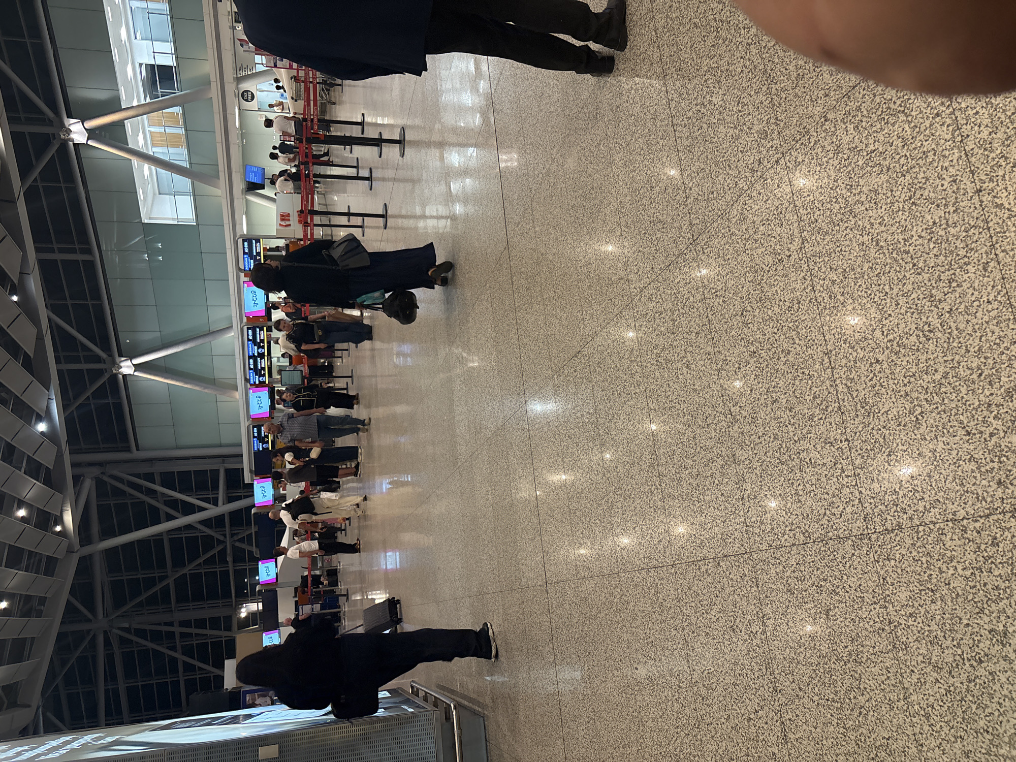 The image depicts a view inside an airport terminal, likely during the daytime given the bright interior lighting. The scene is oriented vertically, presenting a perspective looking down a long row of check-in counters.
The primary subjects are people, observed in various poses within queues and at the counters. Several individuals are seen standing in line, some with luggage, while others are interacting with staff at the check-in desks. The clothing of the individuals is varied, but many are wearing dark colors. One person in the foreground is dressed in black, with a black bag. Further down the line, other passengers are visible, some with carry-on bags and rolling suitcases.
The setting is a modern airport terminal with a polished, speckled floor. Large structural beams and a high ceiling with overhead lighting are prominent features of the architecture. The check-in counters are equipped with digital displays showing information, and red velvet ropes are used to delineate the queues. Some of the digital displays show text in what appears to be Armenian script, possibly indicating flight information or check-in desk assignments. The overall atmosphere appears to be that of a busy, functional travel hub. There are no obvious indicators of weather or specific time of day beyond the indoor illumination. The emotions of the individuals are not discernible due to the distance and image quality.