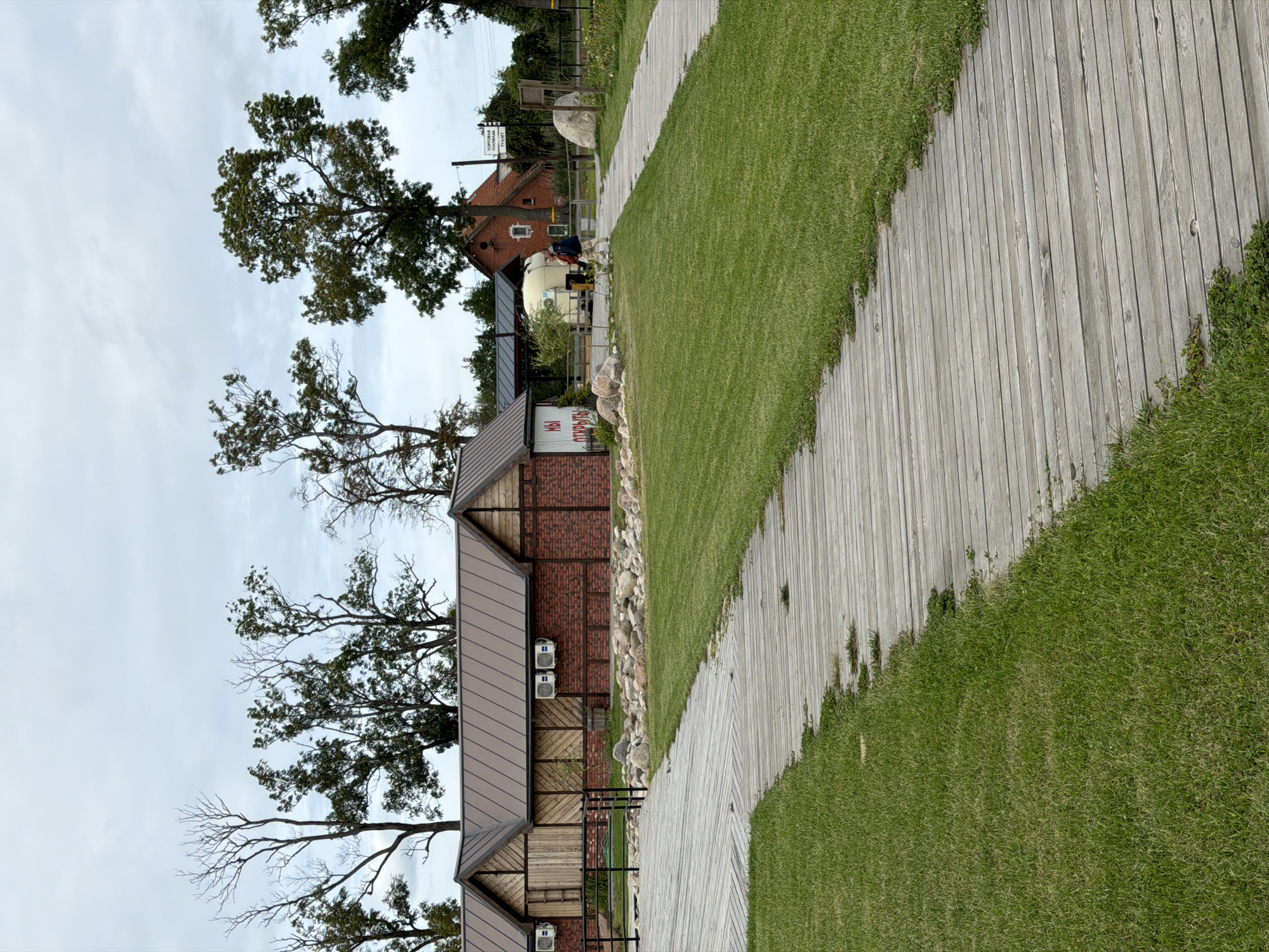 The image depicts a park-like setting in Nekrasovo, Russia, with a focus on two winding wooden boardwalks traversing a lush green lawn. In the background, several structures are visible, including a low, modern building with a brown metal roof and brick and wooden accents, two air conditioning units mounted on its exterior, and a sign with Cyrillic text. Adjacent to this building is a white trailer with a rounded top. Further in the background, a larger, traditional red brick house with a pitched roof and a chimney stands. A small cluster of trees with sparse foliage is situated behind the modern building, while a dense line of trees with fuller canopies is visible further in the distance. A single person, dressed in a blue jacket and light-colored pants, is walking on the right boardwalk. The sky is overcast with a pale grey hue.