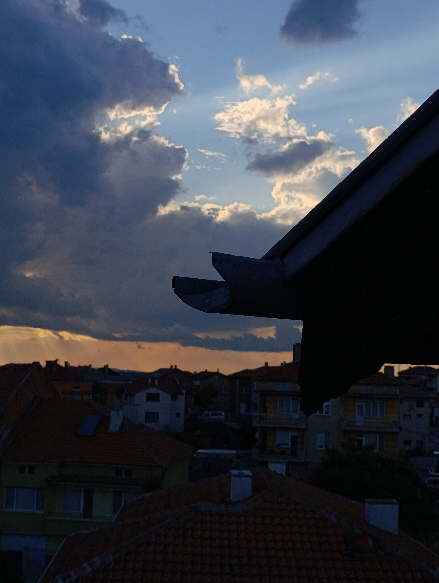 The image depicts a rooftop view in Svilengrad, Bulgaria, under a dramatic sky. Thick, dark clouds dominate the left side of the frame, while patches of bright blue sky break through, illuminated by sun rays, toward the center and right. The orange glow of a setting sun is visible along the horizon line. In the foreground, a dark roof and guttering system, out of focus, obstruct part of the view. Below, a cluster of residential buildings with red tile roofs is visible, ranging from single-story to multi-story structures. One building has a blue solar panel on its roof. The overall impression is of a twilight scene after a storm.