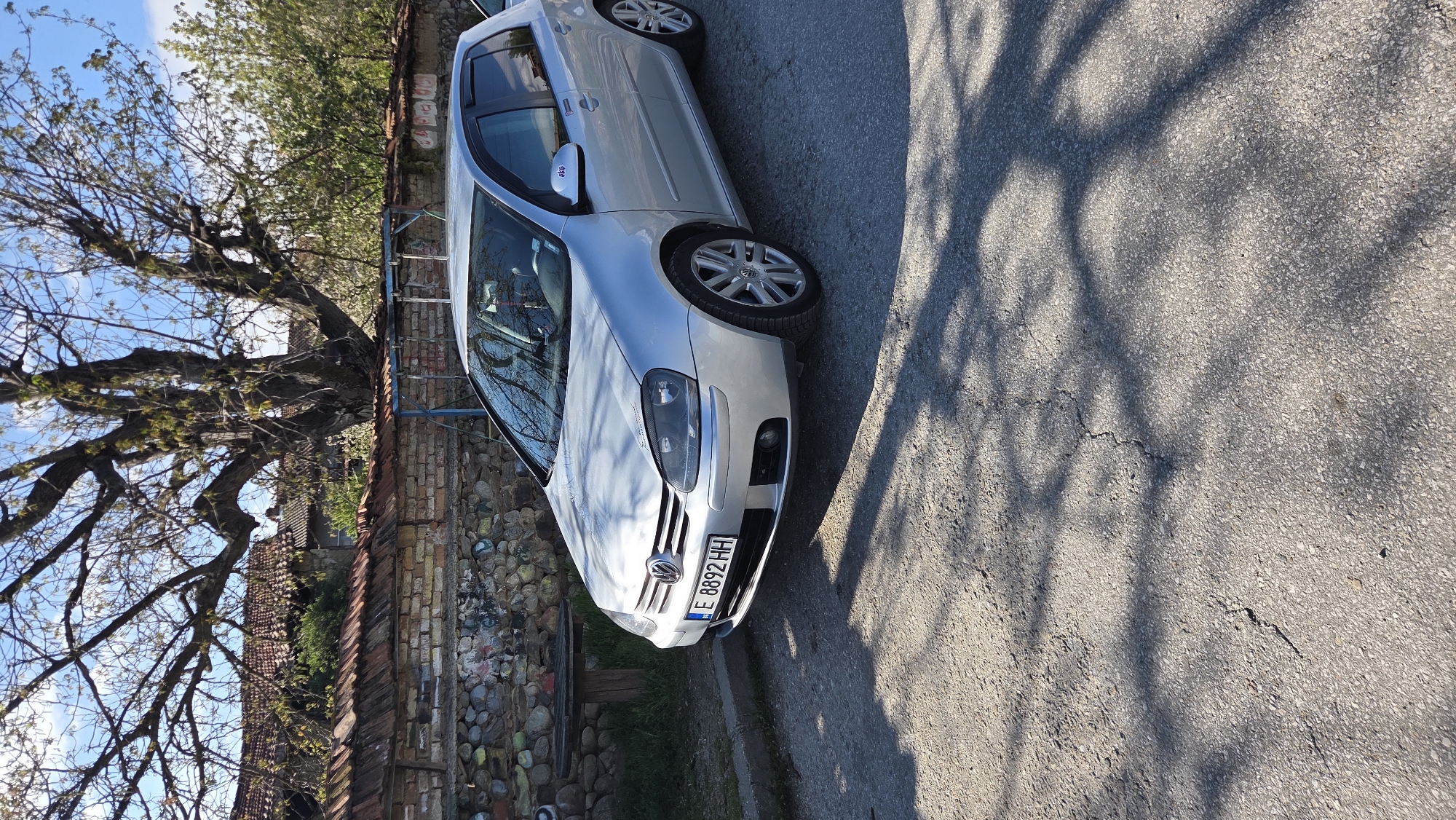 The scene is an outdoor shot of a silver Volkswagen Golf parked on an asphalt street in Unknown, Unknown. The car is angled slightly towards the camera, revealing its front grill, headlights, and front passenger side. The license plate reads "E 8892 HH." To the left of the car is a weathered brick and stone wall, next to a tree with bare branches that stretch towards the sky. Shadows cast by the tree are visible on the asphalt street. The sky is blue with some scattered clouds.