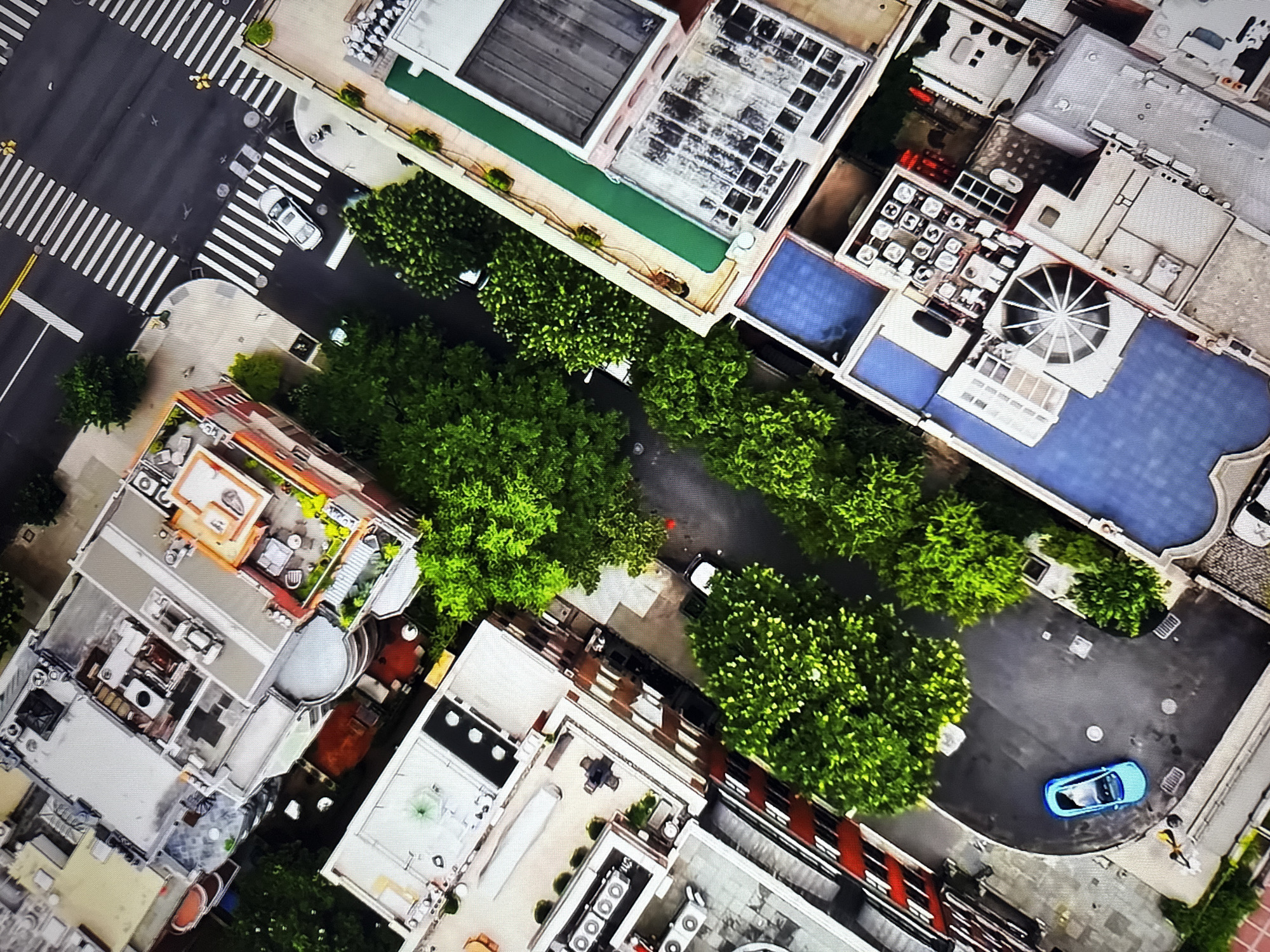 An aerial view shows a city intersection with cars and buildings. In the foreground, a blue car is parked in a curved driveway next to lush green trees. To the right, a building has a blue tiled roof and a circular skylight. In the background, a silver car is stopped at a crosswalk. Buildings of various heights and architectural styles line the streets, with some featuring rooftop terraces and trees. The overall scene depicts a moderately dense urban environment with a mix of residential and commercial structures, interspersed with significant green spaces. This image is not located in Burgas, Bulgaria.