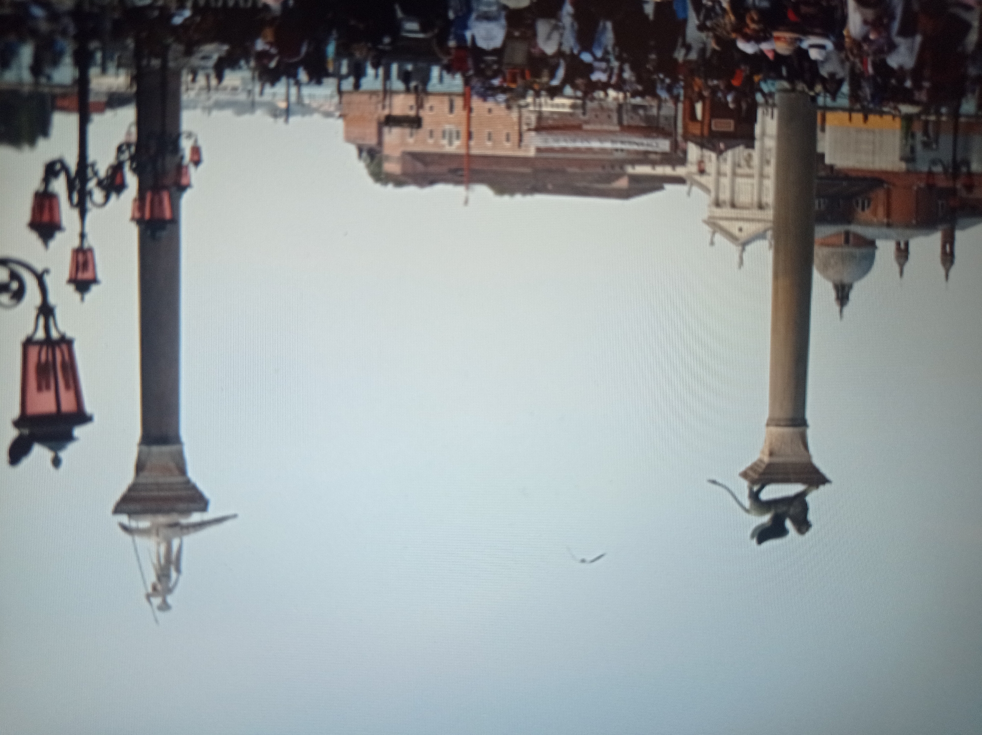 The image depicts a wide view of a public square with numerous people milling about. Dominating the scene are two tall columns, each topped with a statue. The column on the left features a winged lion, while the column on the right is surmounted by a figure holding a spear. In the background, architectural structures with domes and facades are visible, suggesting a historical urban setting. The sky is a light blue, with a faint bird flying in the distance. Several lampposts, some with multiple lights, are also present. The overall impression is of a bustling gathering in an open-air location. The scene is not in Mukachevo, Ukraine; it is Saint Mark's Square in Venice, Italy, famous for these very columns and statues.