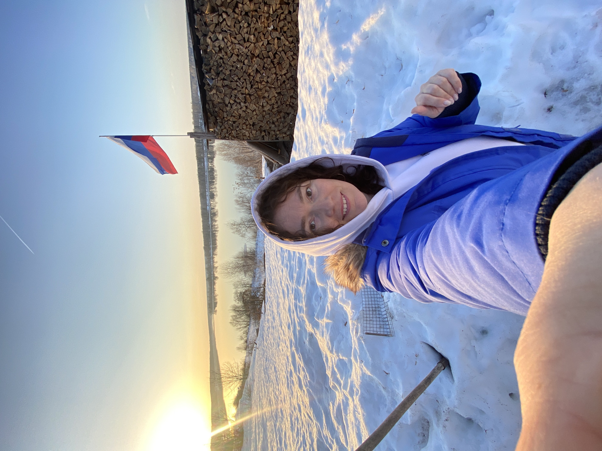 This image depicts an outdoor scene in Ольховка, Russia, during what appears to be either sunrise or sunset, characterized by warm, golden light. The foreground is covered in textured snow, reflecting the light. A woman, approximately mid-20s to 30s, occupies the center-left, taking a selfie. She has dark, curly hair and is wearing a blue winter jacket over a white hooded sweatshirt, with the white hood covering part of her hair. She is smiling faintly and looking directly into the camera. Her right arm is extended, holding the camera, with her hand visible in the lower-left corner. Her left hand is partially visible near her midsection. To her left, a wooden handle or stick is partially embedded in the snow, and a metal grid object containing small snow or ice lumps rests on the snow. In the midground, a wide body of water (likely a river or large lake) spans the horizon, reflecting the bright sky. The far bank is lined with bare trees. To the right of the woman, a tall stack of neatly piled firewood is visible, reaching a height comparable to the woman. Above and slightly to the left of the firewood stack, a Russian flag, displaying white, blue, and red horizontal stripes, flies on a pole against a clear, gradient sky that transitions from bright yellow-orange near the horizon to light blue overhead. A faint white contrail is visible in the upper-central sky.