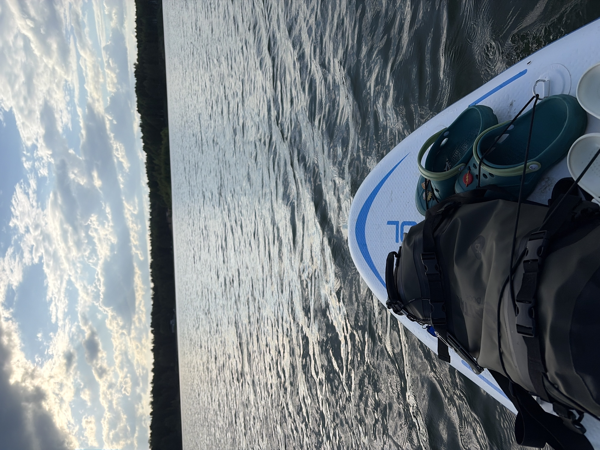 The image presents a view from a paddleboard on a body of water, likely a lake or river. The paddleboard is white with a blue stripe. On the board are a pair of green Crocs sandals and a dark-colored dry bag secured with black straps and buckles. The water exhibits gentle ripples and wavelets, reflecting the sunlight. In the background, a strip of land covered in dense trees is visible beneath a sky filled with scattered clouds. The overall setting suggests a calm, outdoor recreational activity. There is no information available in the image to suggest that this is located in Selychka, Russia.