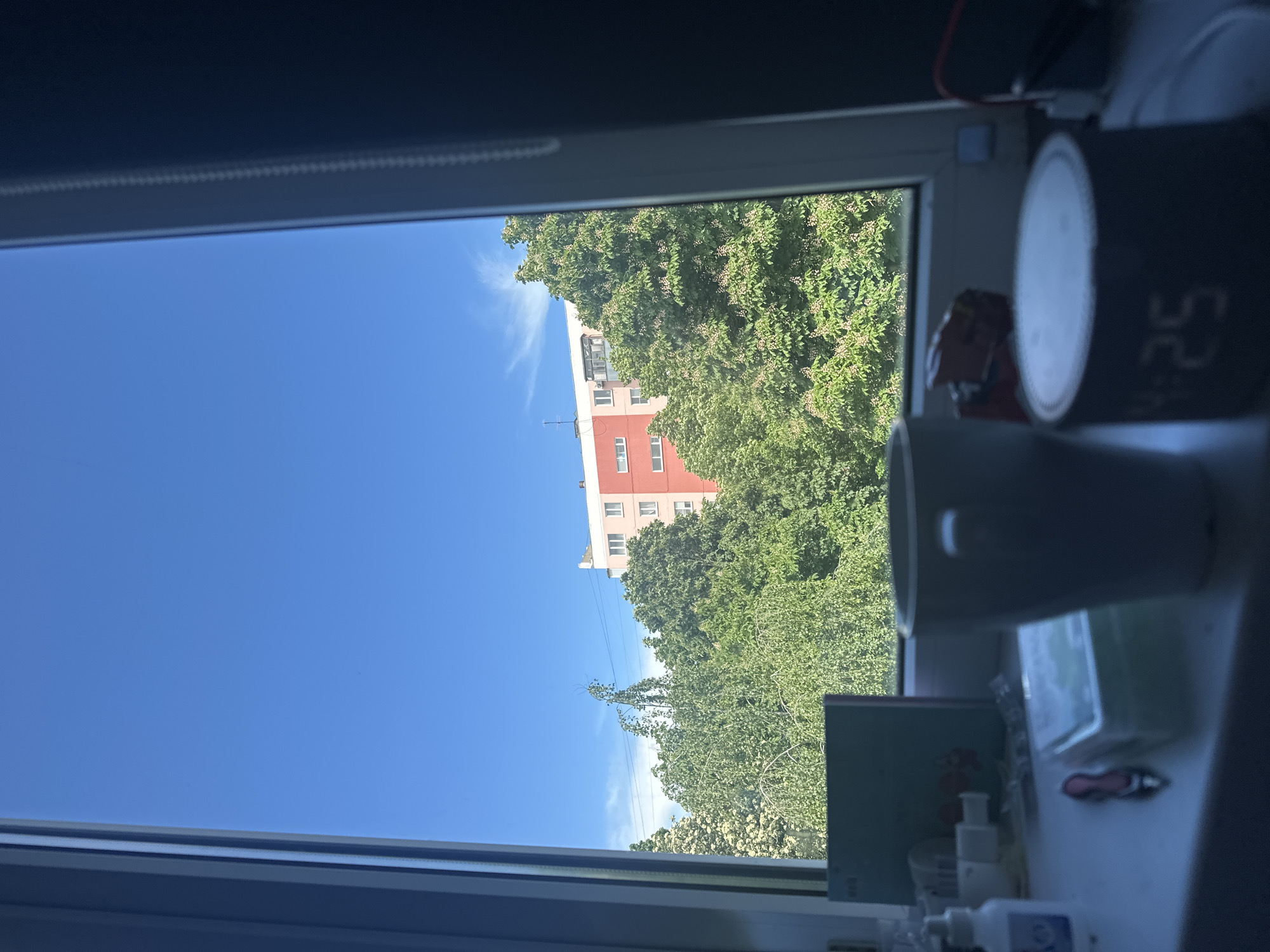 From the vantage point of a window, a view outside includes a cloudless blue sky partially obscured by lush green trees. A multi-story building with red and white sections is visible through the foliage. On the windowsill, various objects are present, including a clock displaying "5:25", a mug, and other miscellaneous items. Given the information in the filename, the location of this scene is Stavropol, Russia.