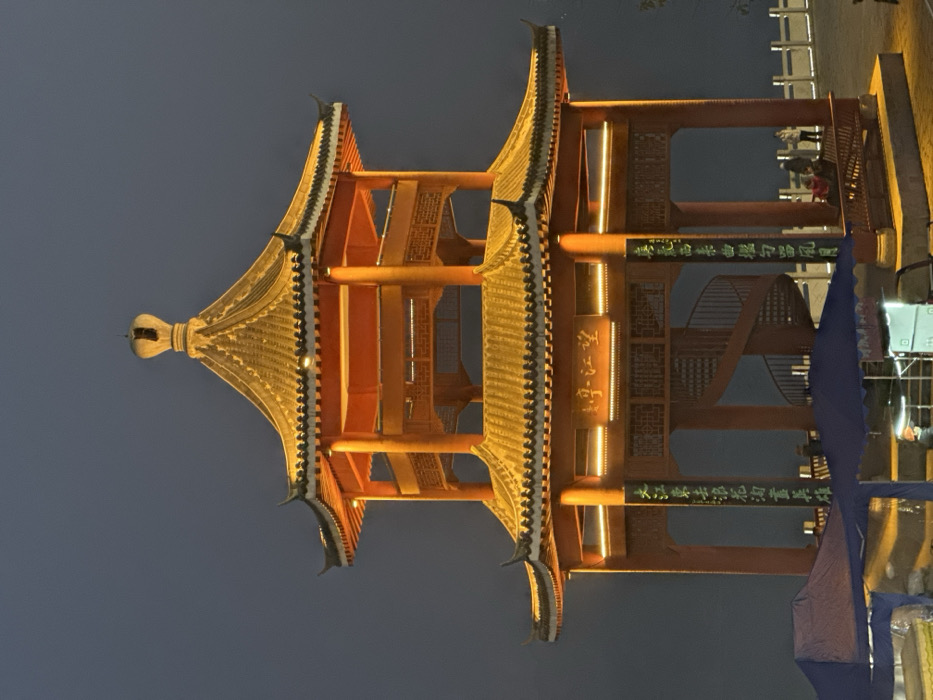 The image displays the illuminated Wangjiang Pavilion (望江亭) at night in Jiangjin, China. The two-tiered traditional Chinese pavilion is constructed primarily of orange-red timber, featuring golden-yellow tiled roofs with upturned eaves and a central finial. Strip lighting, emitting a warm golden glow, is installed beneath the eaves and along the main horizontal beams of both levels. Vertical banners with Chinese characters, including "大江東去浪淘盡" on the left pillar, adorn the support structures. A horizontal plaque above the lower entrance clearly displays the name "望江亭". Inside the pavilion, a dark brown spiral staircase ascends to the upper level. On the lower level, three individuals are seated on a bench to the right, with one additional person standing nearby, all positioned towards a white railing system in the background. In the lower-left foreground, two blue-canopied tents partially obstruct the view of the pavilion's base; one tent covers what appears to be a vendor setup, including a white box-shaped object, possibly a speaker. The sky is uniformly dark blue-gray, indicating nighttime conditions.
