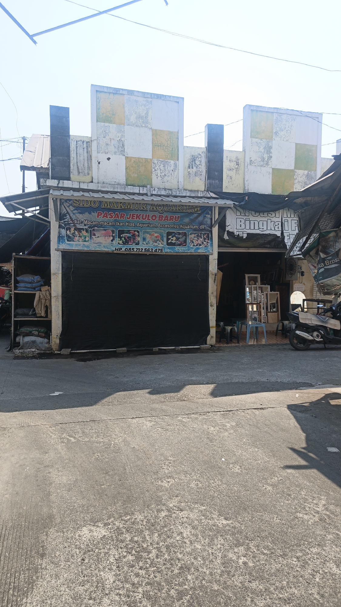 The image shows the exterior of a small shop in Jekulo, Indonesia, identified by a banner reading "SIDO MAKMUR AQUARIUM PASAR JEKULO BARU". The shop appears to be an aquarium store, as indicated by the banner's text "Sedia: Macam-macam Ikan Hias, Aquarium, Acesories Aquarium, dll." (Available: Various Ornamental Fish, Aquariums, Aquarium Accessories, etc.). A black tarp covers the storefront, suggesting it is closed or under renovation. To the left of the entrance, a shelf unit is visible, containing bags of what appear to be sand or substrate. To the right of the entrance, some furniture and framed items are displayed outside, including chairs, a small table, and several framed pictures or mirrors. A motorcycle is parked to the far right. The building itself has a faded facade with a checkerboard pattern of yellow and white squares. The surrounding environment appears to be a street or marketplace, with a concrete road in the foreground and overhead wires suggesting an urban setting.