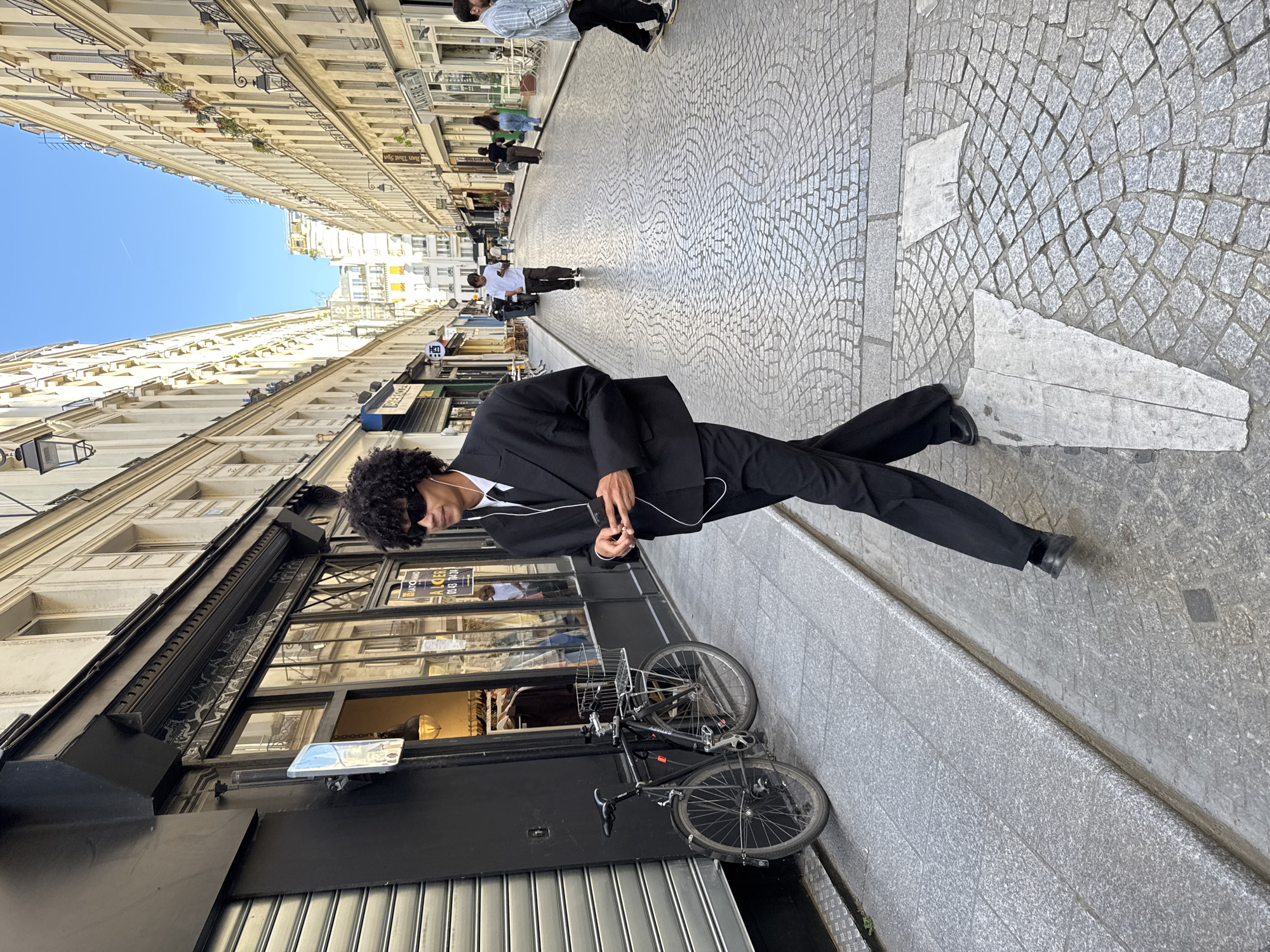 The image captures a vibrant street scene in Paris, France, on a clear, sunny day. The main subject is a young man with dark, curly hair, wearing a black suit, white shirt, and dark sunglasses. He has wired earbuds in his ears and is holding a smartphone in his right hand, appearing to be walking or posing on the cobblestone sidewalk.
The street is lined on both sides by classic Parisian multi-story buildings, constructed from light-colored stone with numerous windows and some balconies. The ground is paved with intricate cobblestones. A black bicycle is parked next to the building where the main subject is standing. In the background, other pedestrians are visible further down the street. The sky above is a bright, clear blue.
Visible text includes a "A LOUER" (For Rent) sign with a phone number (01 83 72 84 24) on a shop window, and another sign in the distance that appears to read "Ban Thai Spa."