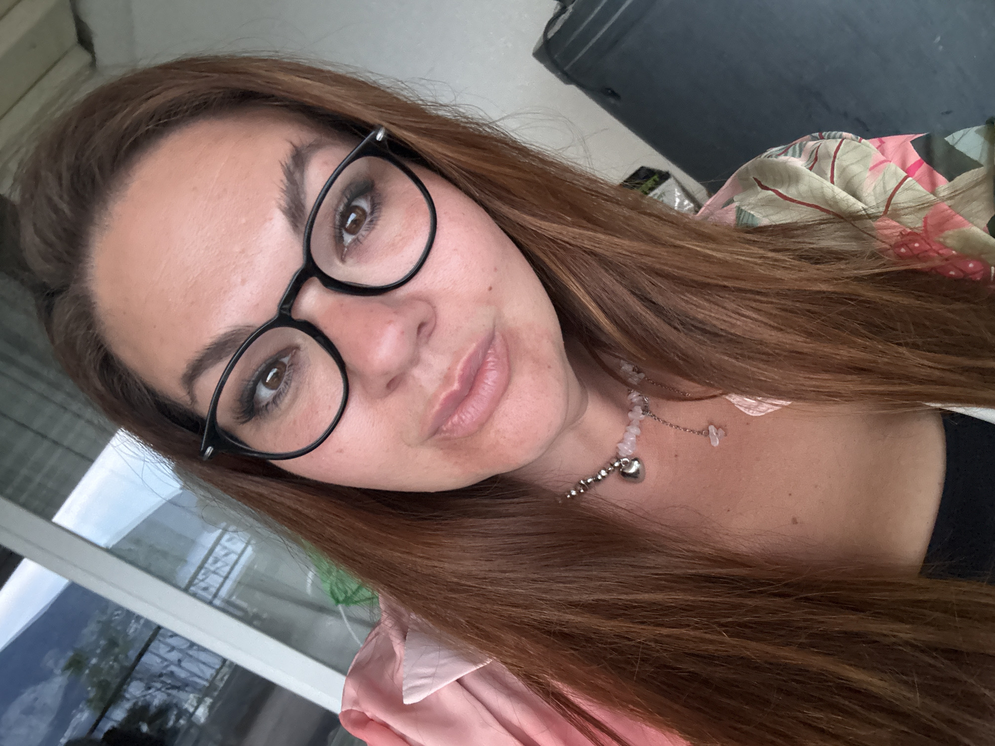 A selfie of a woman with brown hair, wearing black framed glasses and a necklace with a heart pendant. She is wearing a pink shirt. The background shows a window with a reflection of greenery and sky. A piece of fabric is draped in the background. The location cannot be verified based on the image.