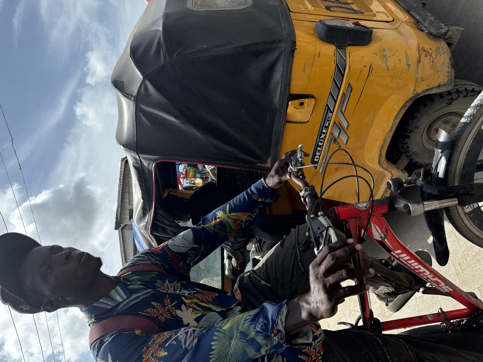 A man in a patterned shirt and black cap is seen working on a red bicycle in Ado, Nigeria. The bicycle is positioned in front of a yellow tricycle, a common mode of transport in the city. The tricycle is labeled "DELUXE PLUS." The sky is visible in the background.