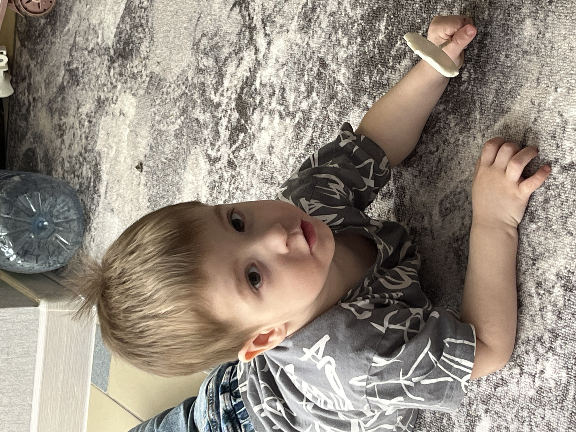 A fair-skinned young boy with short, blonde hair is lying on a gray and white patterned carpet. He is wearing a gray t-shirt with white leaf-like designs, and the top of denim pants is visible near his waist. He is looking up towards the viewer with a slight pout. His right arm is extended, and he is holding a piece of white, cylindrical material between his index and middle fingers. A large, clear plastic water bottle is visible in the upper left corner of the image. A gray and pink toy is in the very top left corner of the image. The exact location of the image within Chaikovsky, Russia, is not evident.