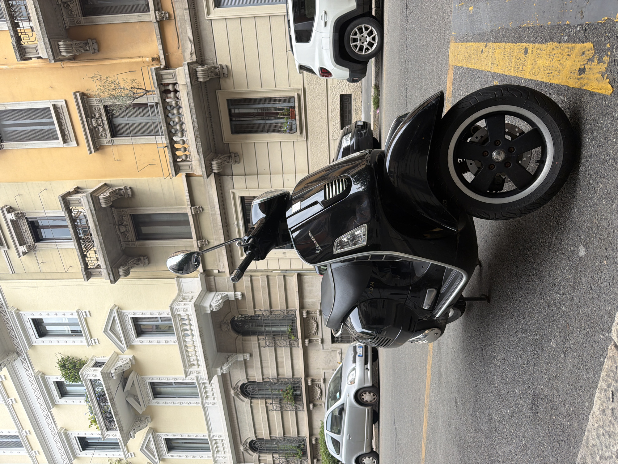 An outdoor, eye-level, medium shot captures a black Vespa scooter parked on an asphalt street in Milan, Italy. The scooter is positioned in the foreground, angled towards the right side of the frame, with its kickstand down. In the background, several buildings showcase classical European architecture with yellow and cream-colored facades, ornate balconies, and detailed window frames. A silver car and a white SUV are parked further down the street, and a yellow painted line is visible on the asphalt next to the scooter. The scene appears to be a typical street in a European city.