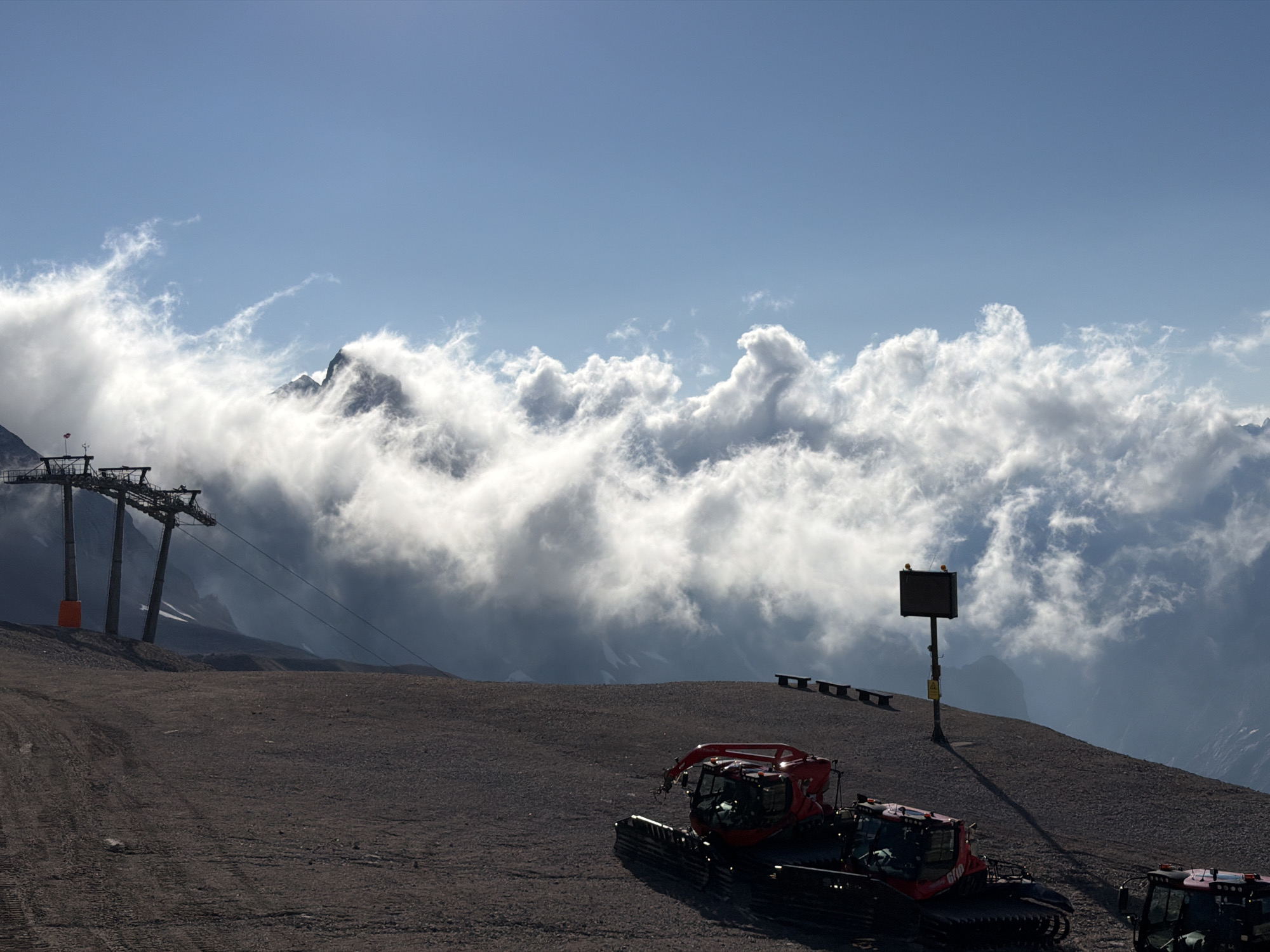 The image depicts a mountainous landscape, likely in the vicinity of Garmisch-Partenkirchen, Germany, given the presence of ski resort infrastructure. In the foreground, two red snow groomers are parked on a gravelly surface. The terrain slopes upwards towards the left, where a ski lift with three pylons is visible, one of which has an orange marker. Beyond the ski lift, a sharp mountain peak pierces through a dense layer of white clouds. The sky above is a clear blue, with some wispy clouds. In the middle ground, a signpost with a digital display stands on the slope, accompanied by several benches. The overall scene suggests a high-altitude ski resort, possibly during the off-season or early morning before operations commence. The lighting indicates a bright day, possibly late morning or early afternoon.