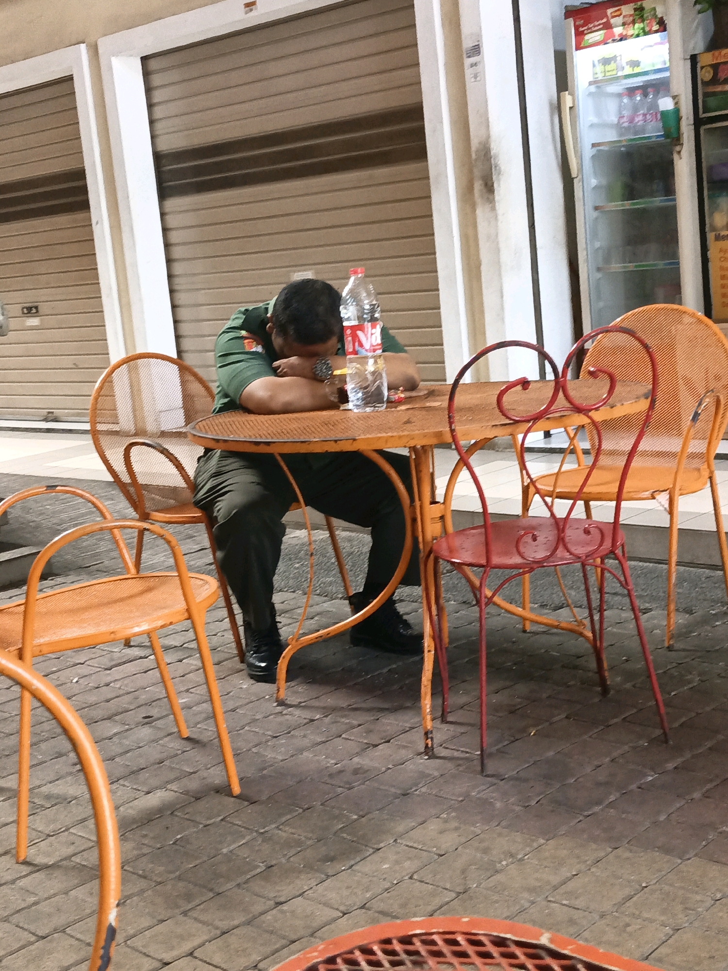 A man in a green uniform is asleep with his head on a table at an outdoor cafe in South Jakarta, Indonesia. He is sitting on an orange chair and his arms are resting on an orange table. A bottle of water is on the table next to him. There are other orange chairs and a red chair around the table. The cafe appears to be located in front of a building with closed metal roll-up doors. The ground is paved with brick-like stones. The lighting suggests it is evening or night.
