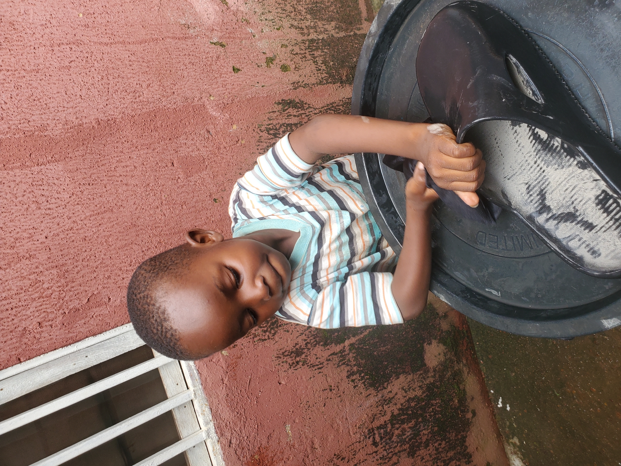A young boy, likely of African descent, is the sole subject in this horizontal, outdoor photograph. He is wearing a striped t-shirt with horizontal bands of white, teal, orange, and black. The boy is positioned next to a large, black, plastic basin, which takes up a significant portion of the right side of the frame. His hands are inside the basin, suggesting he is interacting with something within it, possibly washing or playing. He is leaning forward, his eyes are closed, and he has a slight smile on his face, conveying a sense of contentment or enjoyment.
The background consists of a reddish-brown, textured surface, possibly a wall or ground, with some areas of dampness and mossy growth, indicating it might have rained recently or the area is prone to moisture. To the left of the boy, there is a slatted wooden structure, resembling a window or part of a fence, with a mesh screen behind it. The lighting suggests it is daytime, possibly overcast or in the shade, as there are no harsh shadows. The overall scene appears to be in a residential or utilitarian outdoor area in Jos, Nigeria.
A small amount of text is visible on the rim of the black basin, appearing to be the word "LIMITED" in uppercase letters. The focus is primarily on the boy and his engagement with the basin, creating an intimate and candid portrait.