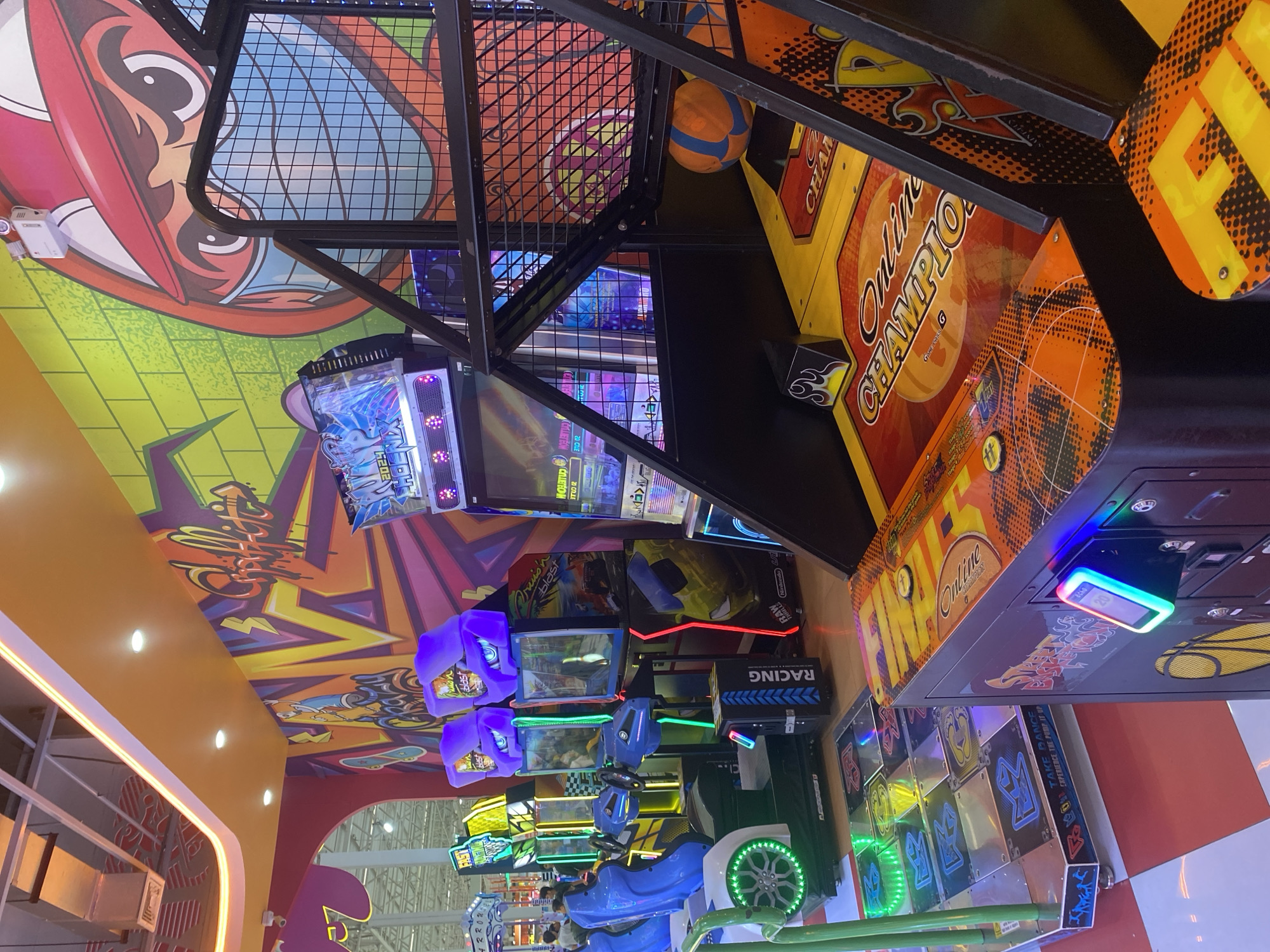 This image captures the interior of an entertainment venue, possibly an arcade, in Si Sunthon, Thailand. The scene is filled with vibrant colors and patterns on the ceiling, which features animated characters and designs. Multiple arcade games are visible, including a basketball shooting game labeled "Online Champion," a racing game with blue car seats, and a dance pad game. A basketball is suspended near the ceiling. The games are arranged in rows, suggesting a structured layout within the arcade. Bright fluorescent lights illuminate the space, creating a dynamic and energetic atmosphere.