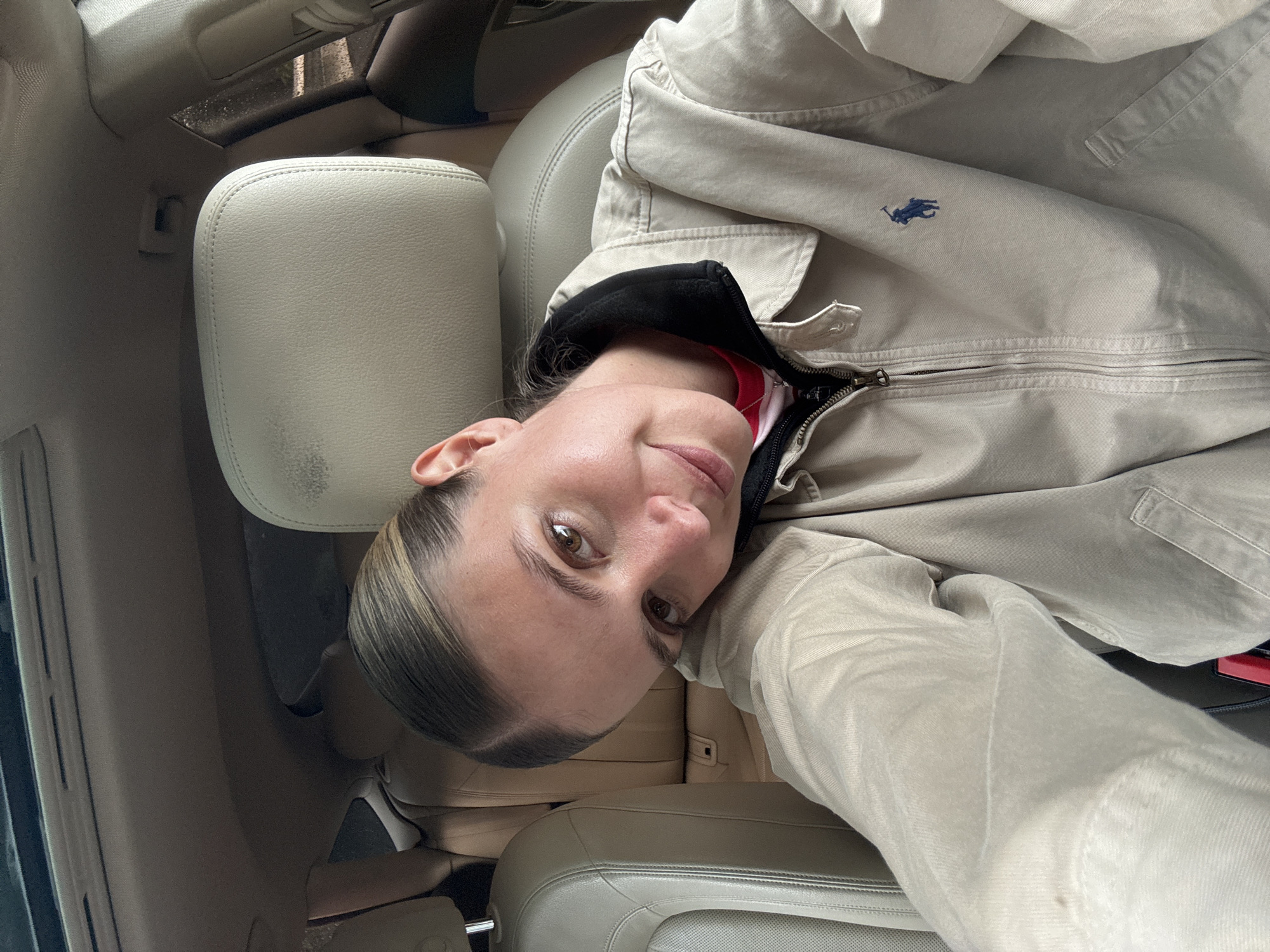 A fair-skinned woman is taking a selfie inside a car. She has light brown eyes and her hair is pulled back. She is wearing a tan Ralph Lauren jacket with a black collar and a hint of a red shirt underneath. The interior of the car has beige leather seats and trim. The exact location in Tbilisi, Georgia, is not discernible from the image.
