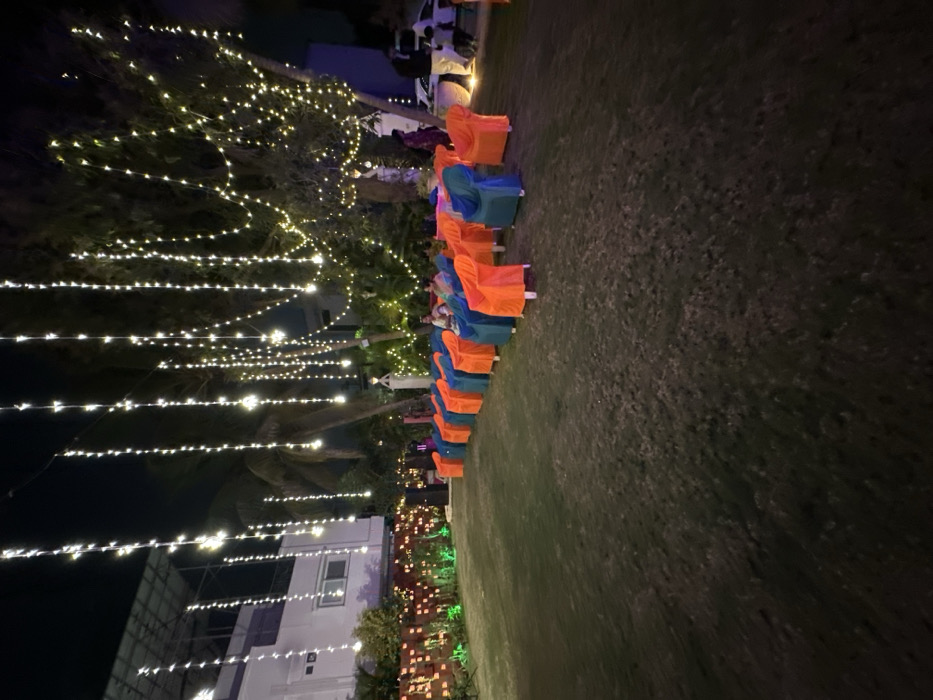 The image captures an outdoor evening event setup on a grassy lawn in Vadodara, India. Numerous string lights, featuring small, warm-toned bulbs, are draped vertically and horizontally across several large trees and extending towards a multi-story white building positioned on the left side of the frame. Two rows of chairs are arranged on the lawn, oriented to face generally left, with each chair covered in alternating bright orange and teal/blue fabric. At least three individuals are visible seated in the second row of chairs. Additional figures are discernible standing in the far right background, near a parked white vehicle. A low hedge or wall in front of the building is decorated with small orange and green lights. The ground is dark green grass, consistent with nighttime illumination.