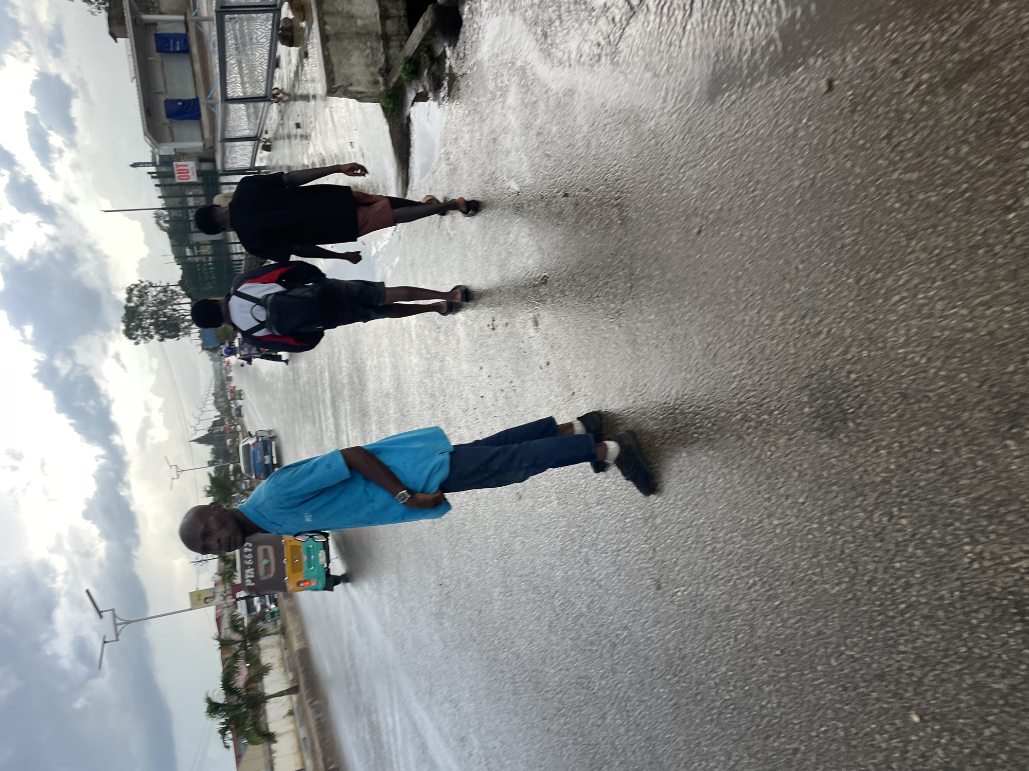 The image depicts a street scene in Barakin Lerawi, Nigeria, under overcast skies, possibly after rain, as the asphalt road surface is wet and reflective. In the foreground, a man wearing a bright turquoise polo shirt and dark pants stands facing left, looking back towards the camera. He has a shaved head and appears to be wearing a wristwatch. Behind him, two other individuals are walking away from the camera down the wet road. The person on the left is wearing a jacket with white and red accents and a dark backpack, while the person on the right is wearing a black t-shirt and reddish shorts. A three-wheeled auto-rickshaw is visible in the background, driving on the wet road. Palm trees line the left side of the road, and buildings, including one with "OUT" signage, are visible on the right. Streetlights are present along the road. The overall impression is of a moderately busy urban street during daylight hours, with evidence of recent precipitation.