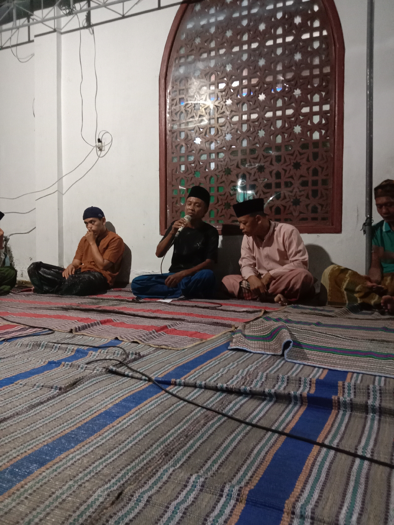 In Brangsong, Indonesia, a group of men are seated on woven mats inside a building. The men are dressed in traditional Islamic attire, including peci caps. One man in the center is speaking into a microphone. The room has white walls and a large, ornate window with a geometric design. The floor is covered with colorful, striped mats, and there are electrical cords visible on the floor and wall. The lighting suggests it is nighttime or an indoor setting with artificial light.
