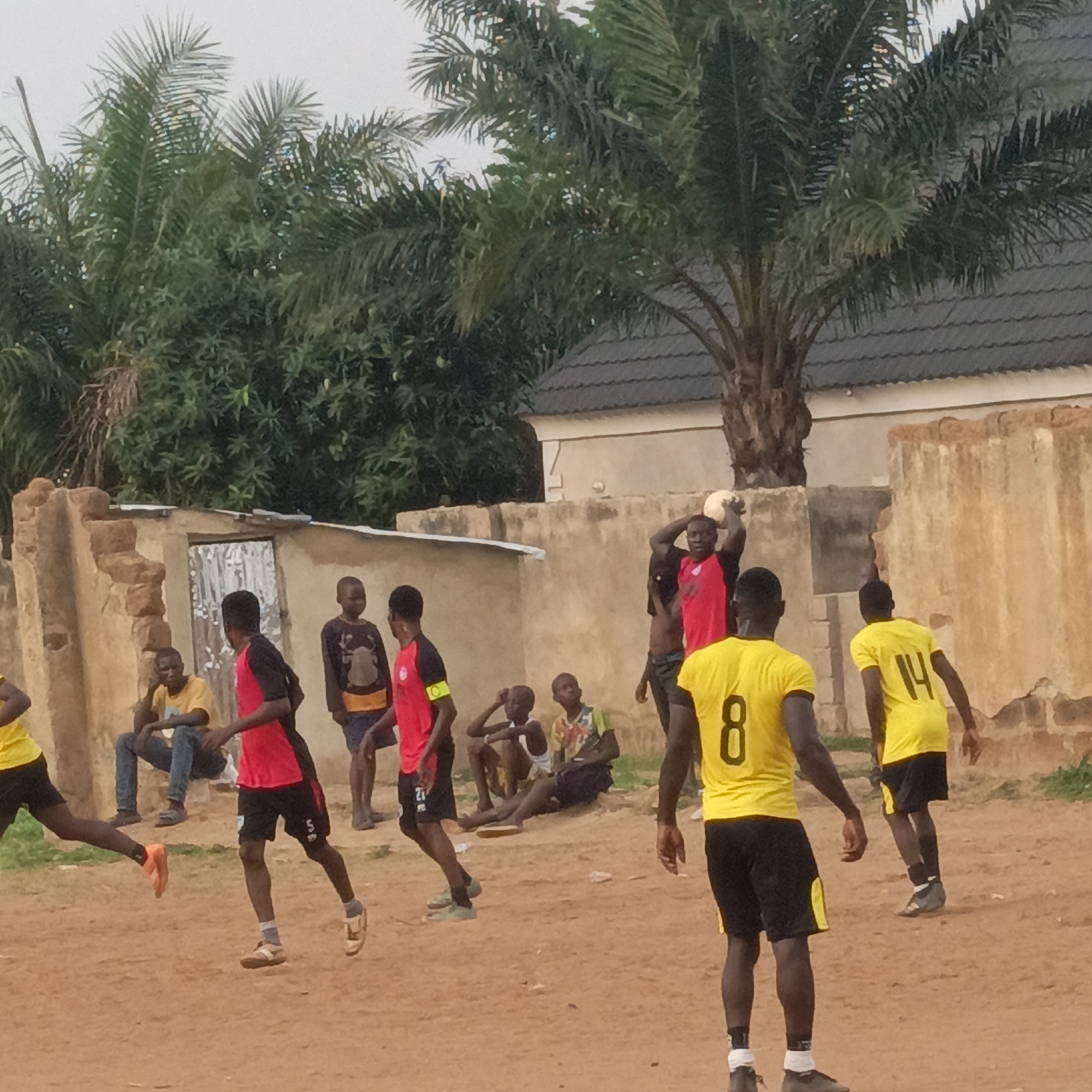 This image depicts a casual outdoor soccer match in Akwanga, Nigeria, featuring several young men and boys. In the foreground and midground, players are actively engaged in the game. A prominent figure in a red and black jersey is preparing for a throw-in, holding a white soccer ball above his head. Two other players in yellow jerseys are visible from behind, one displaying the number "8" and another "14" on their backs. Another player in a red and black jersey wears a yellow identification armband on his left arm, and the letter "S" is visible on his shorts. Footwear among players varies, including sandals, sneakers, and possibly bare feet.
The game is being played on a dry, reddish-brown dirt field. In the background, a few other individuals, likely spectators or resting players, are seated or standing against a low, weathered mud-brick wall. Behind this wall, simple structures, including one with a corrugated iron door, are visible. The scene is framed by dense green foliage and several tall palm trees under an overcast sky. The diffused light suggests it is daytime, possibly late afternoon. The overall atmosphere is one of informal community sport in a rural or semi-urban setting.