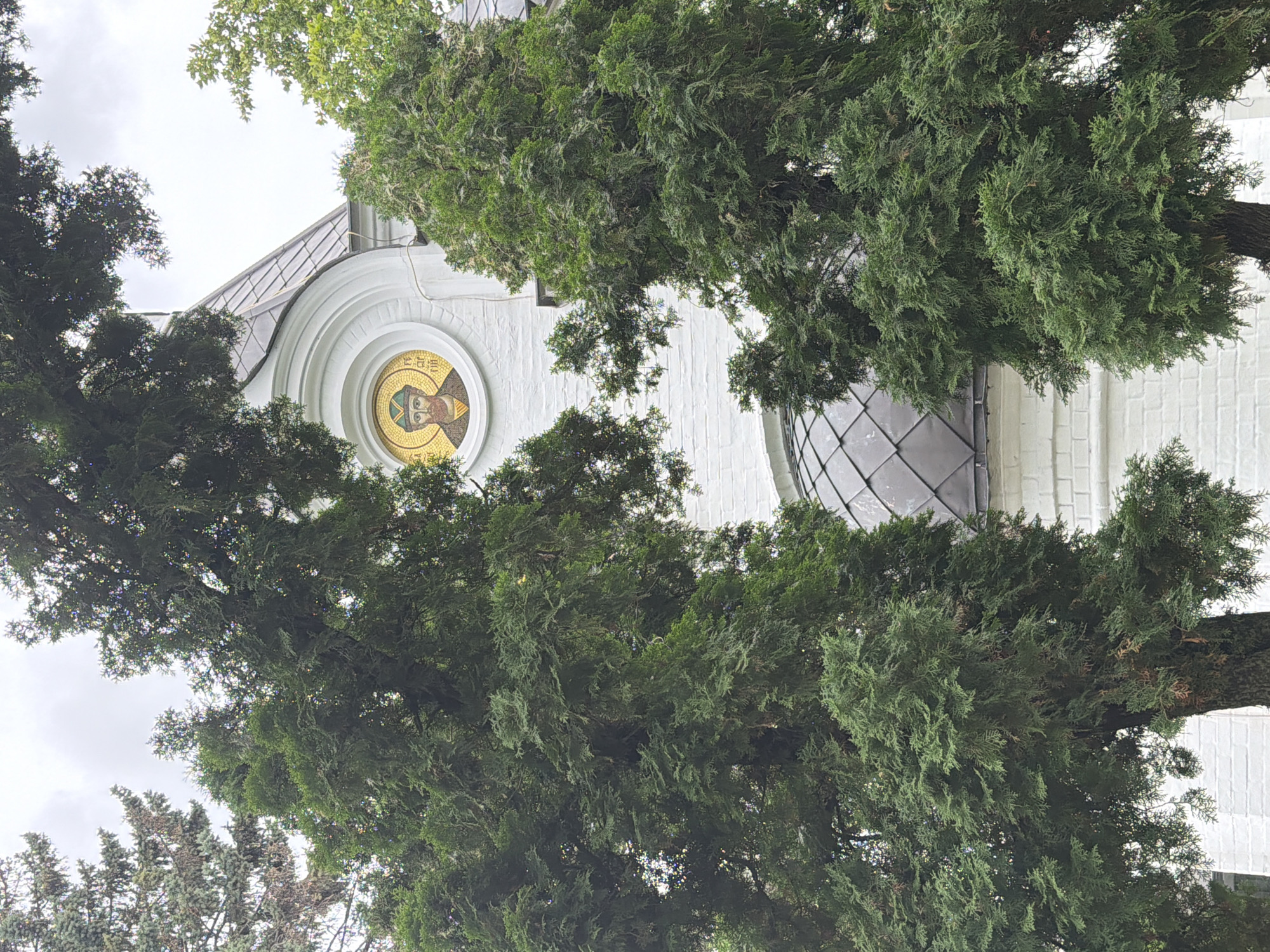 The image shows a close-up view of a white brick building, likely a church, with a dark, sloped roof. A circular mosaic depicting a bearded man in religious attire is visible on the building's facade. Two large evergreen trees with dense foliage partially obscure the building, framing the view from the front. The sky is overcast, suggesting a cloudy day. The building's architecture, including the mosaic, is characteristic of Orthodox churches found in Nizhny Novgorod, Russia. The scene is peaceful and serene, with the natural elements of the trees contrasting with the man-made structure.