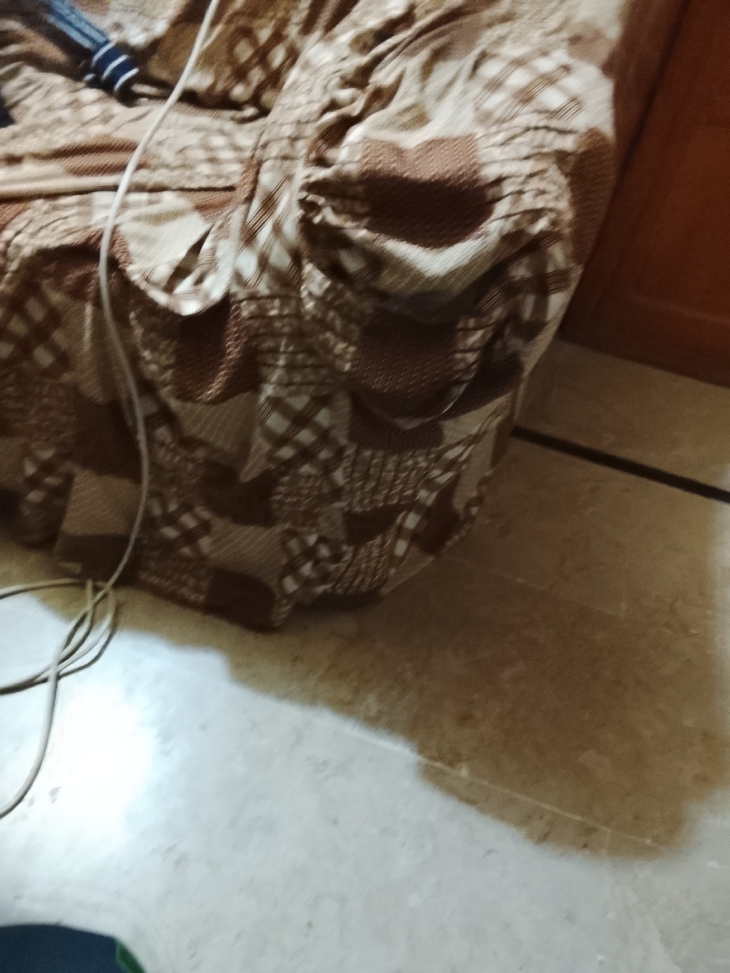 The image shows a piece of furniture covered with a brown patterned cloth. A white cable runs across the cloth and onto the floor. The floor is light-colored tile. The photo was likely taken indoors near a wooden cabinet. No specific information indicates this is in Multan, Pakistan. There are no people present.
