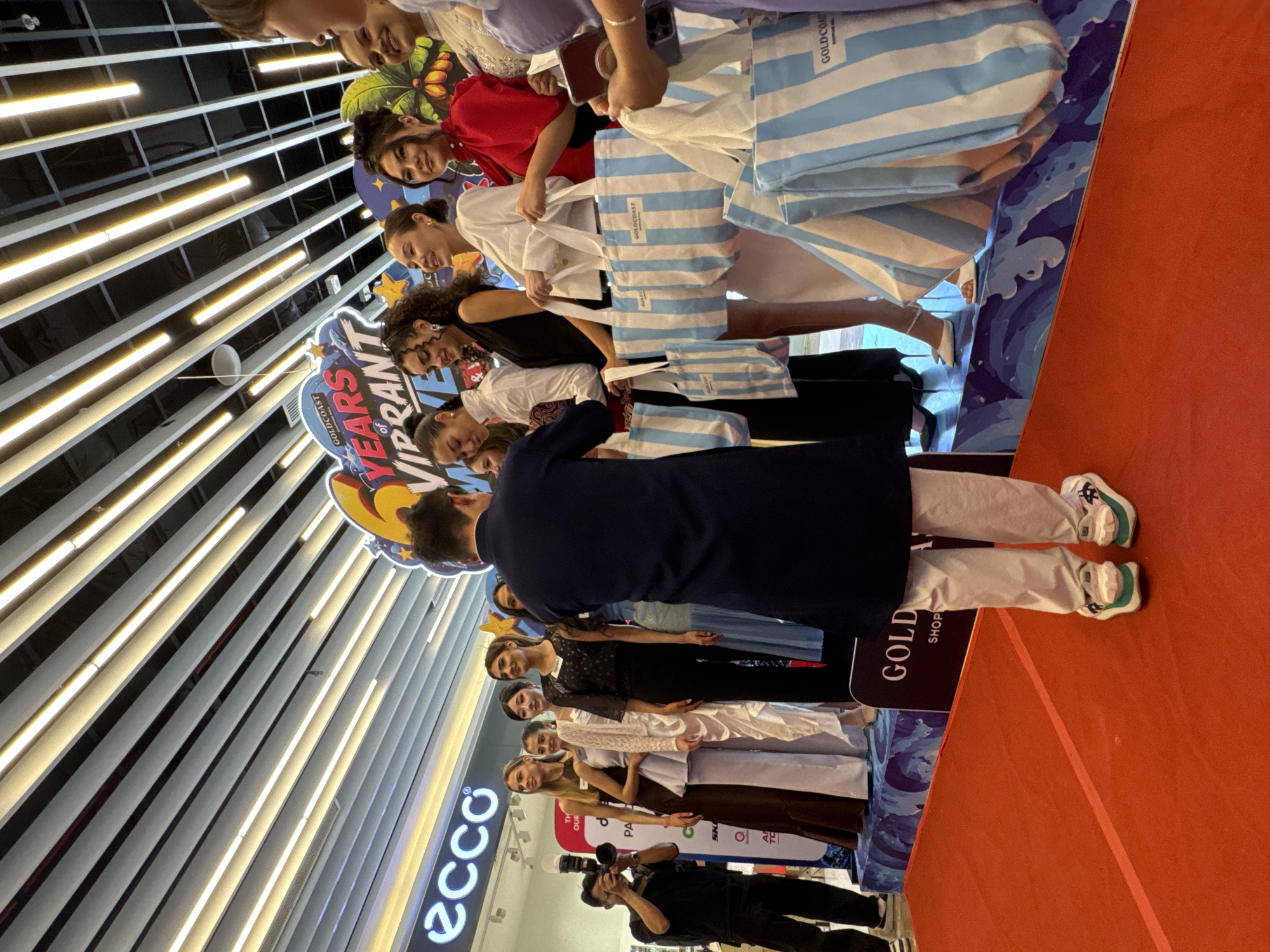 The image depicts an event likely taking place in a shopping mall or public venue, given the presence of store signage (ECCO) and what appears to be a red carpet leading to a raised platform. A group of women, dressed in various formal outfits, are standing on the platform, some holding striped tote bags. A man in a dark tunic and light-colored pants is standing in front of them, facing away from the camera, possibly interacting with the women or presenting something. Another man is visible to the left, operating a camera. The background features a large banner with text including "GOLDCOAST" and "6 YEARS OF VIBRANT." The ceiling has a distinctive design with parallel lines and lights. The overall atmosphere suggests a promotional event or a fashion show. While the prompt mentions Nha Trang, Vietnam, there are no specific visual cues within the image that definitively confirm this location.